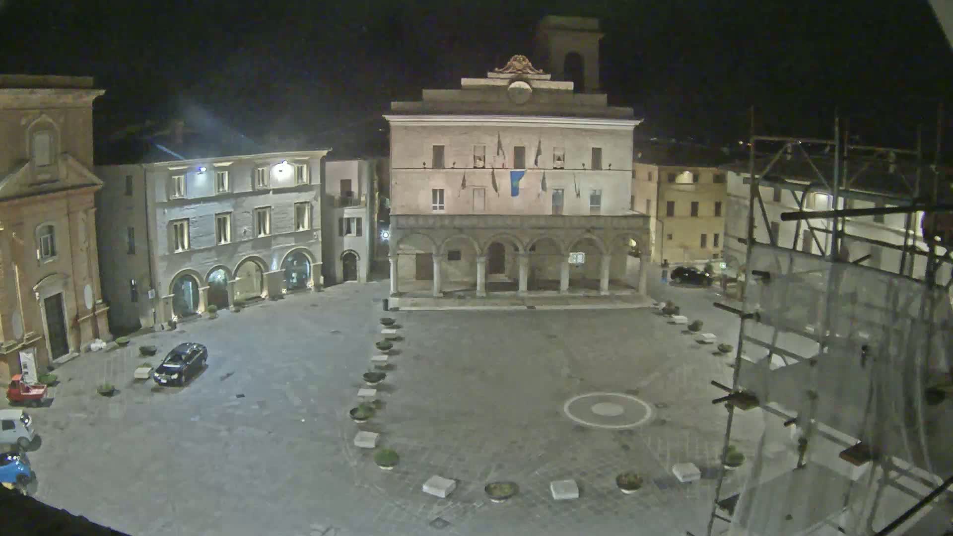 A nighttime view of a town square with several buildings surrounding it, including a large building with arches, under a dark sky.