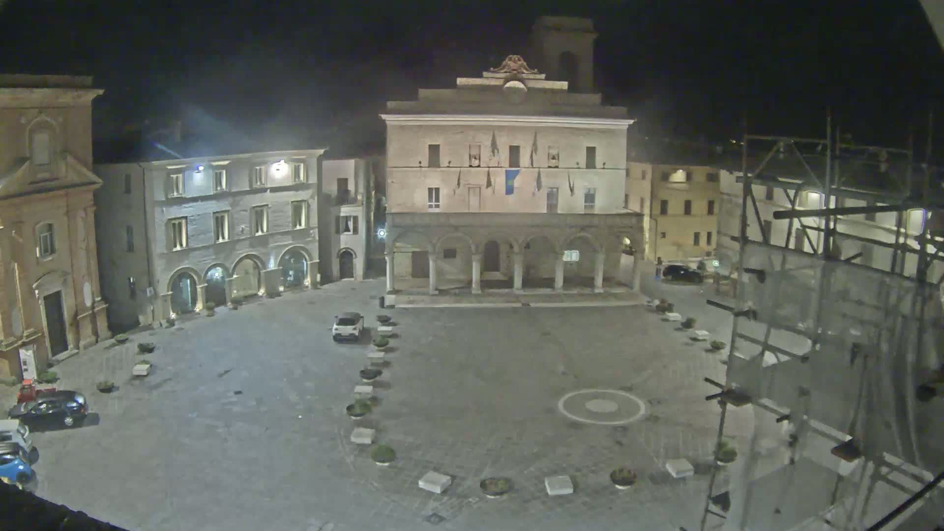A nighttime view of a town square with several buildings, including a large building with a colonnade, and a few cars parked in the square.