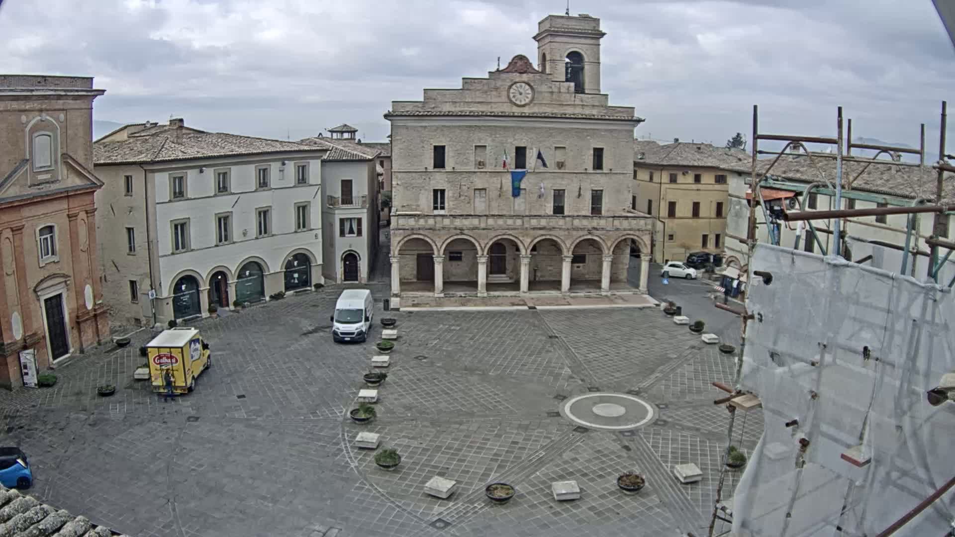 A town square with several buildings, including a large stone structure with arched entryways, under an overcast sky.