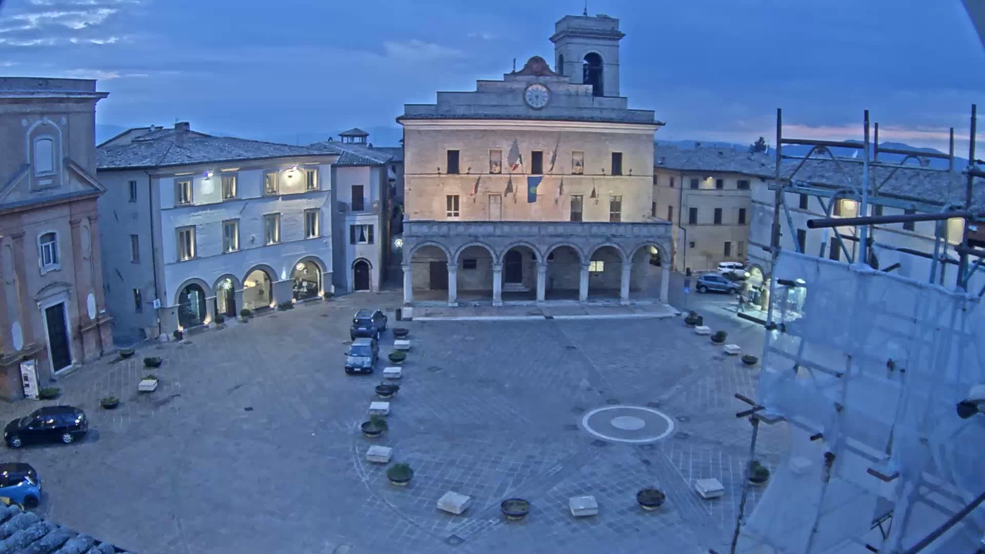 A town square at dusk is surrounded by historical buildings, some with arched entryways, and contains a few parked cars.
