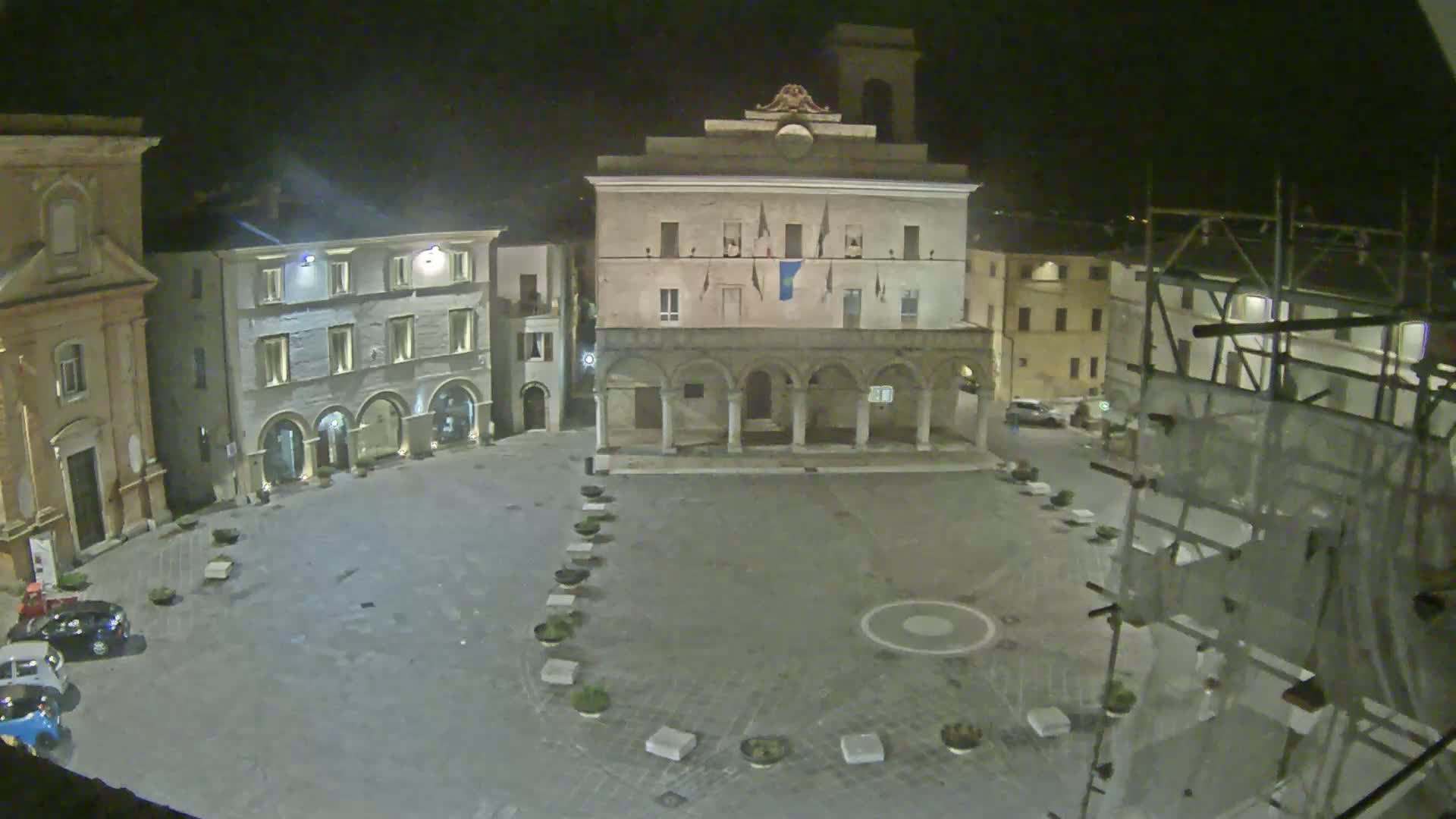 A nighttime view of an empty town square, featuring a large building with arches, other buildings surrounding the square, and some scaffolding to the right.