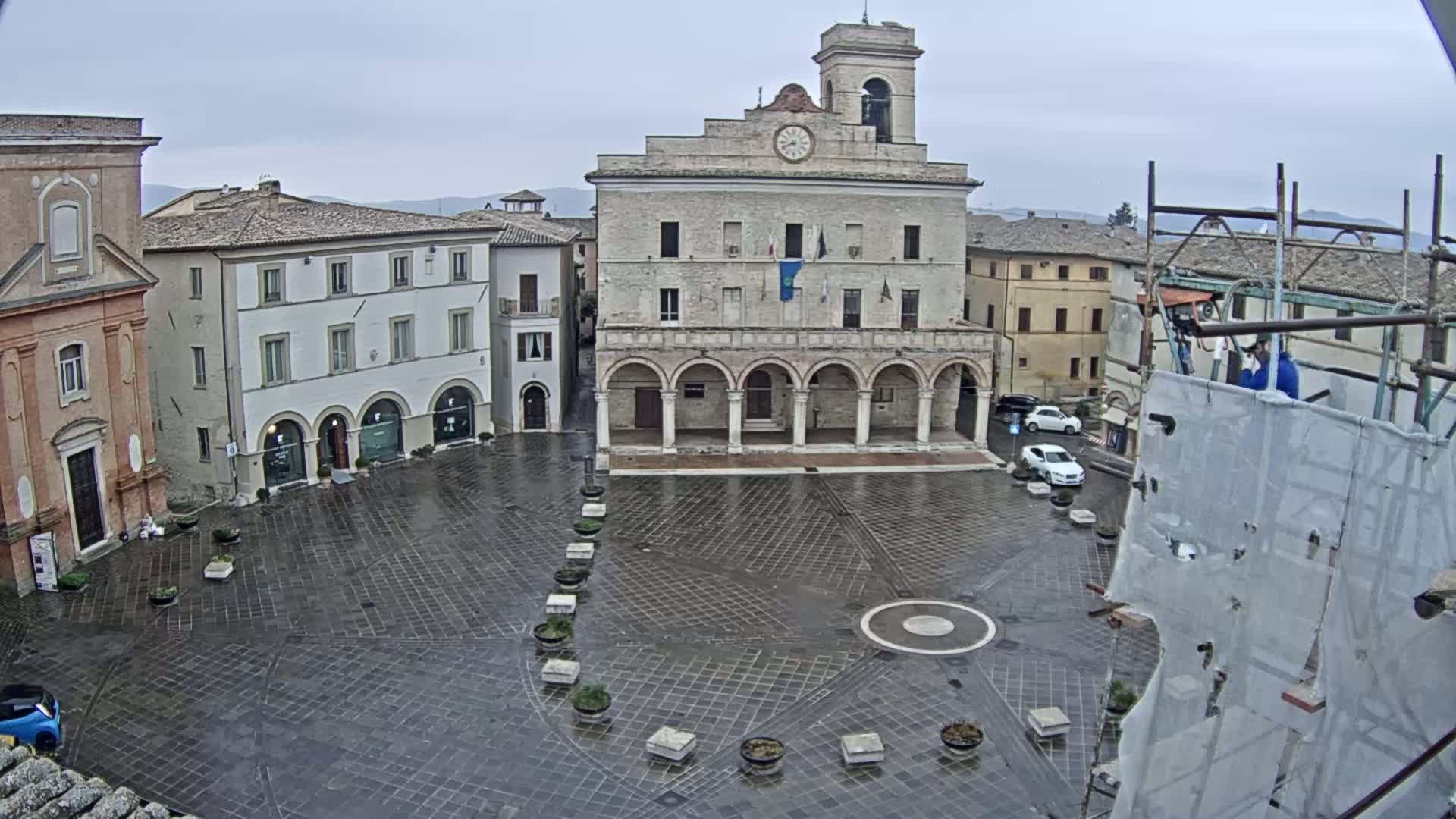 A wet, paved town square is centered on a large, arched building with a clock tower, surrounded by other buildings under an overcast sky.