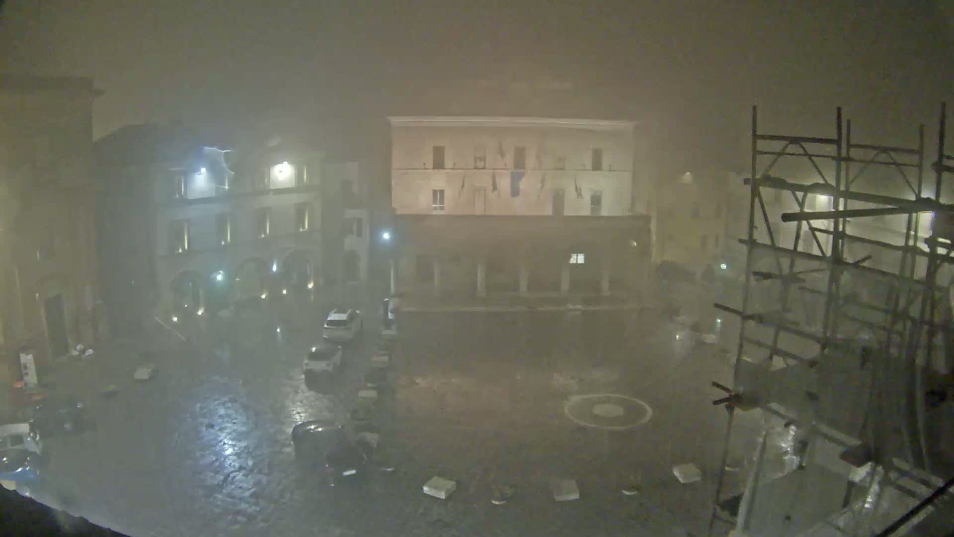 A rain-soaked town square at night is shrouded in heavy fog, with several cars parked and scaffolding visible in the corner.