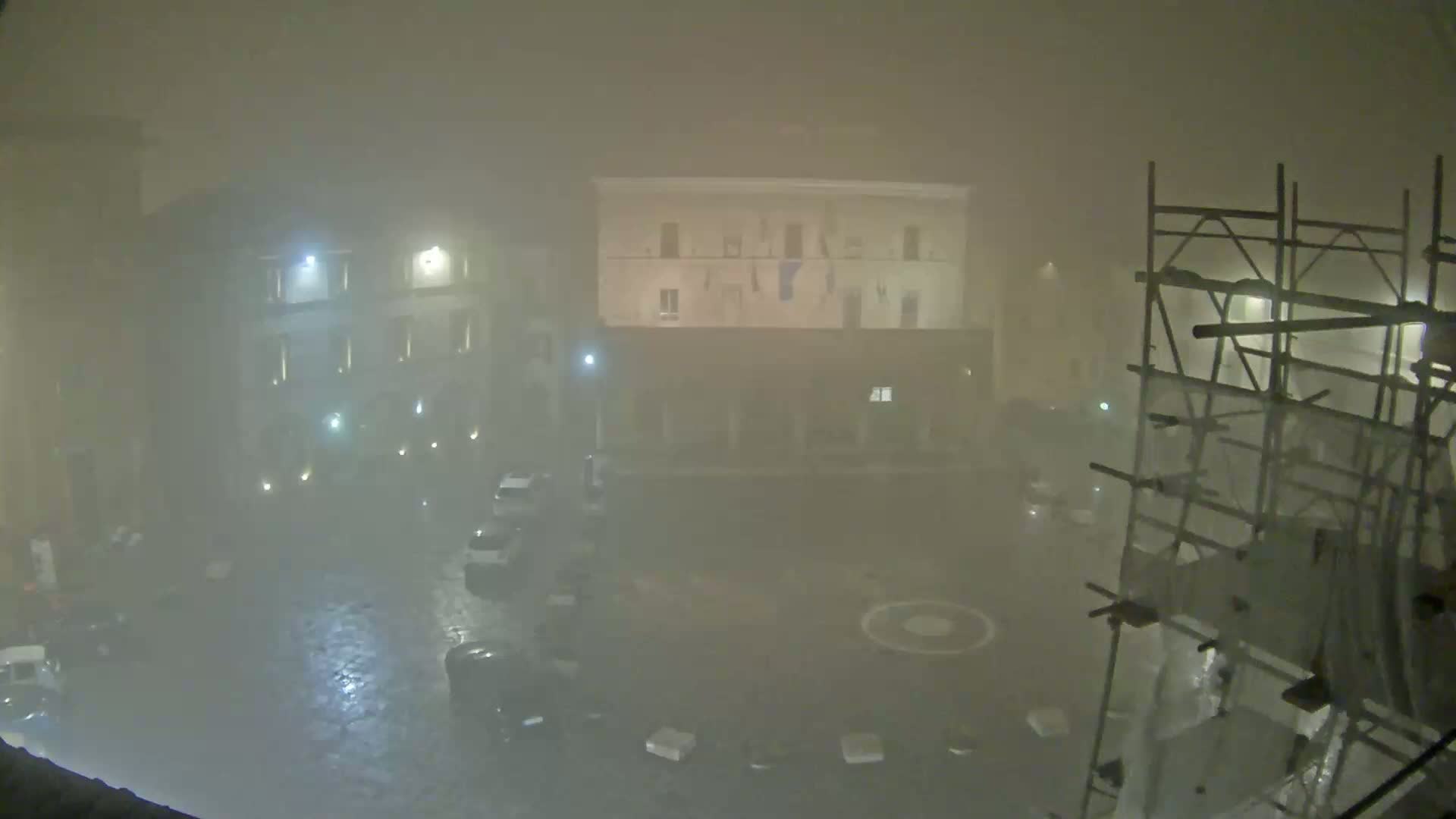 A foggy night scene shows a square with parked cars, buildings, and scaffolding, illuminated by streetlights.