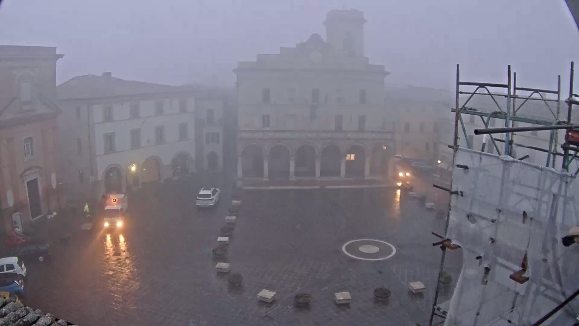 A town square with several buildings is shrouded in heavy fog and rain, with a few vehicles visible in the wet, empty space.