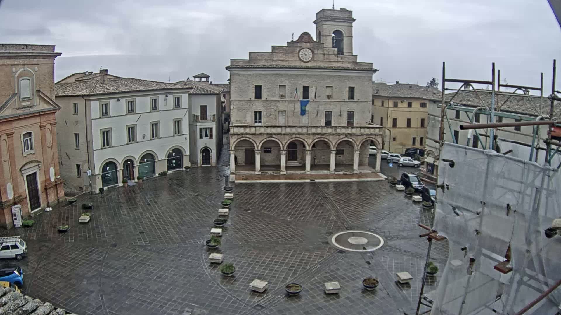 A wet, paved town square is surrounded by historic buildings under a cloudy sky, with scaffolding and construction netting visible in the foreground.