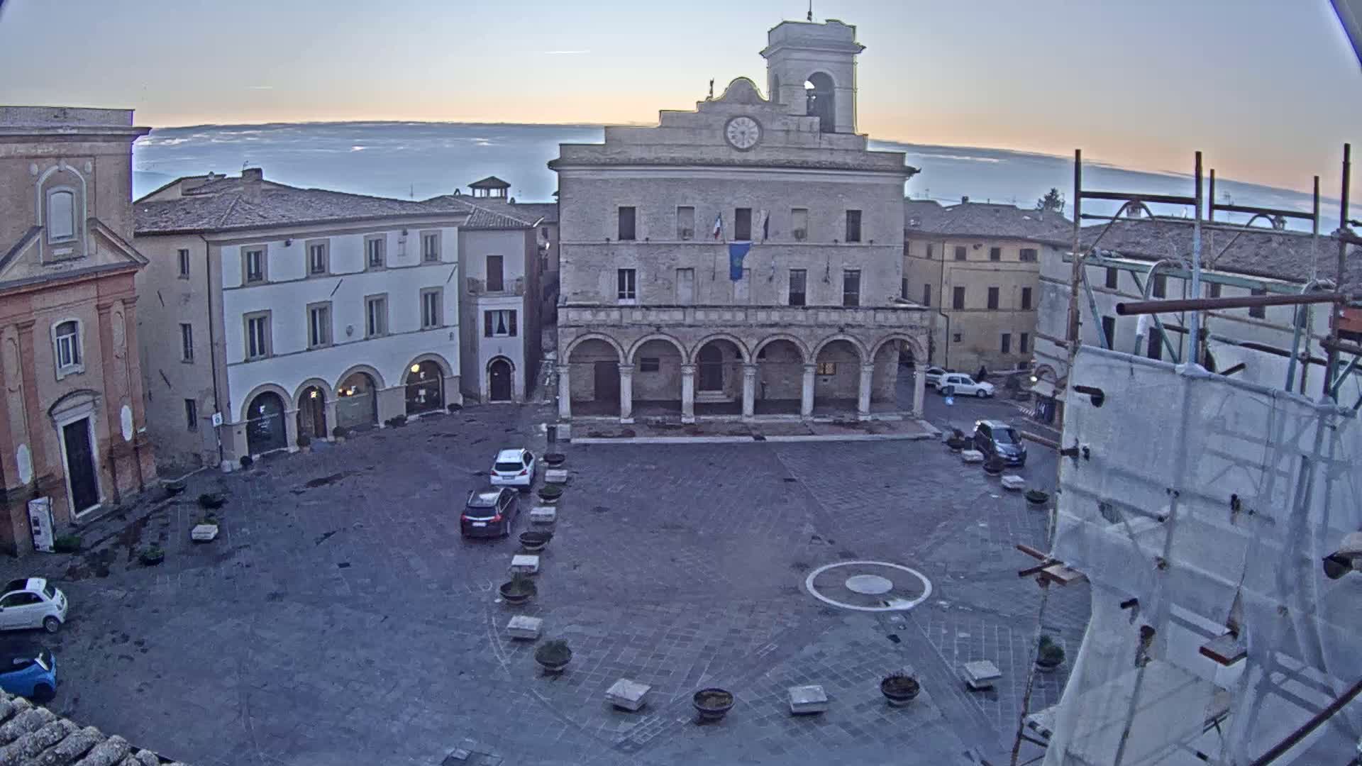 A town square with a large, arched building in the center, surrounded by other buildings, under a mostly clear sky with a hazy horizon.