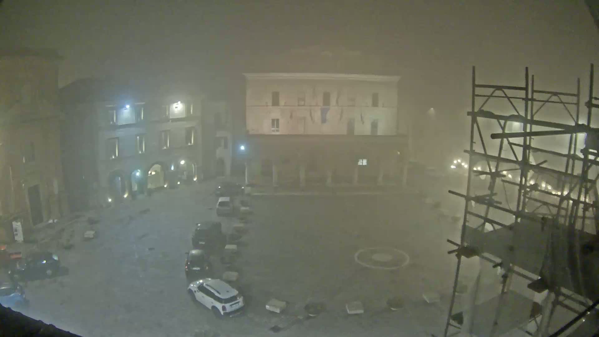 A foggy, nighttime view of a town square with several cars parked and scaffolding in the foreground.