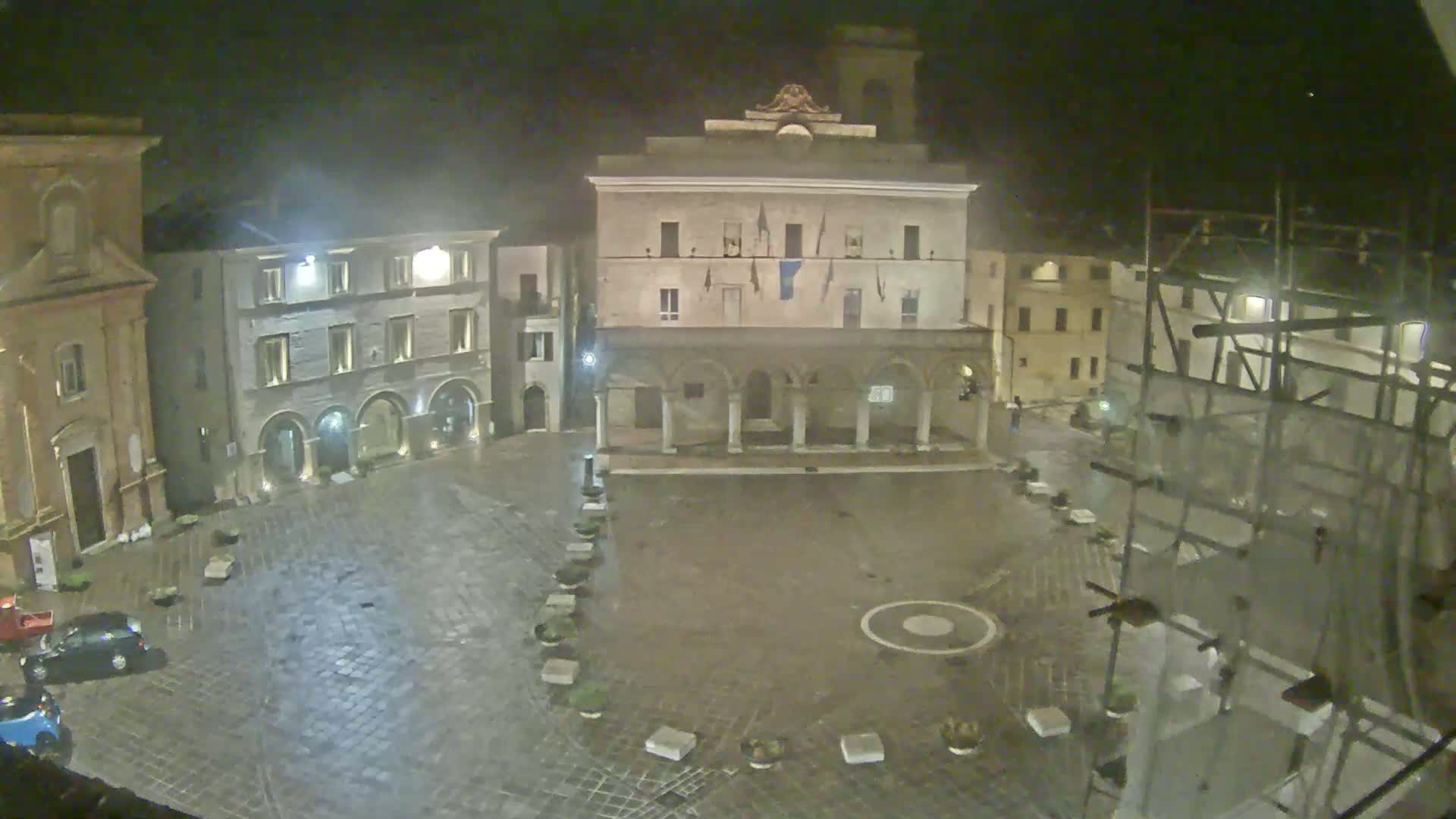 A wet cobblestone town square at night is surrounded by historic buildings, one of which has scaffolding.