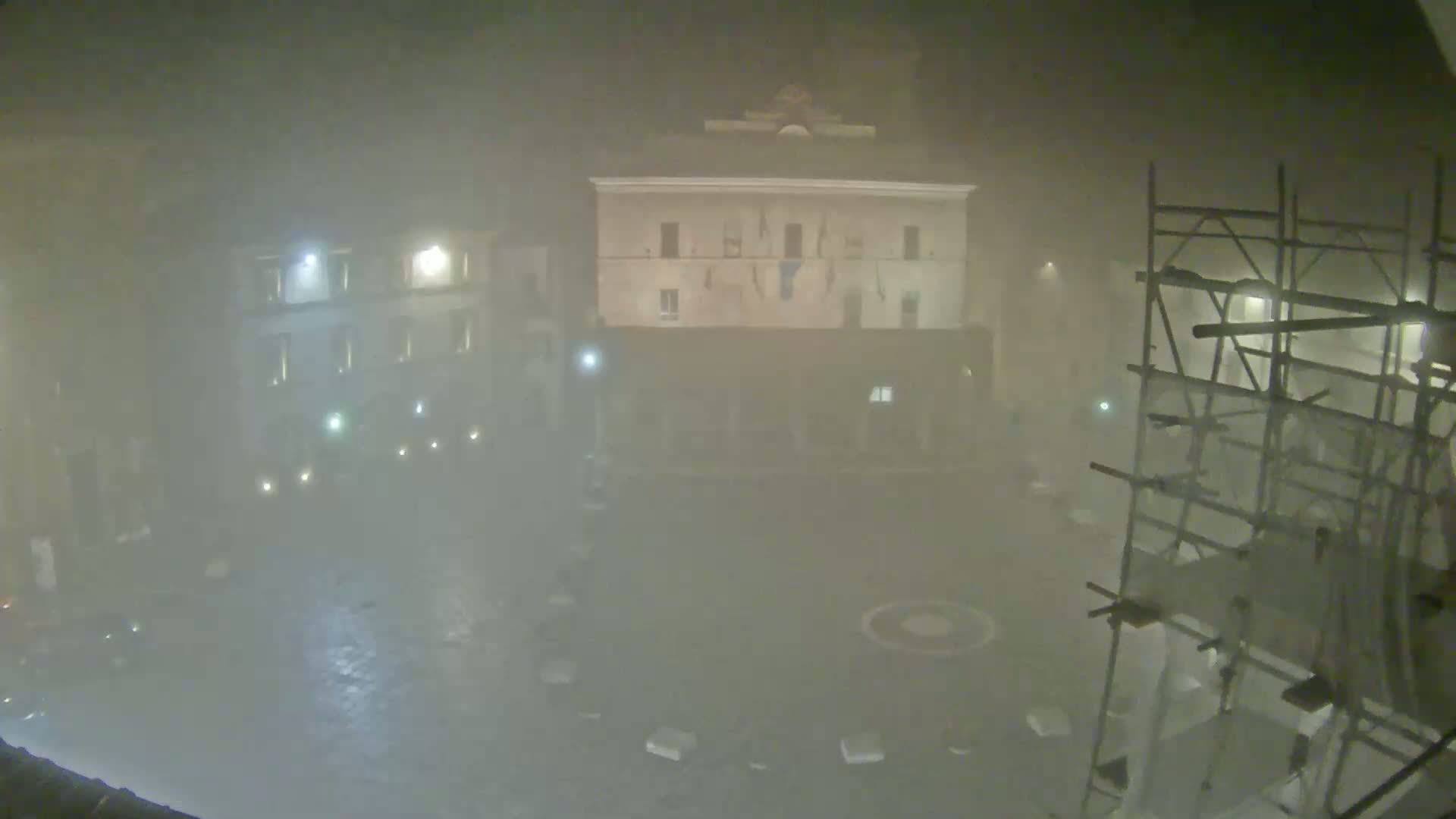 A foggy, nighttime view of a town square with buildings and scaffolding, showing some lights and a wet, dark ground.