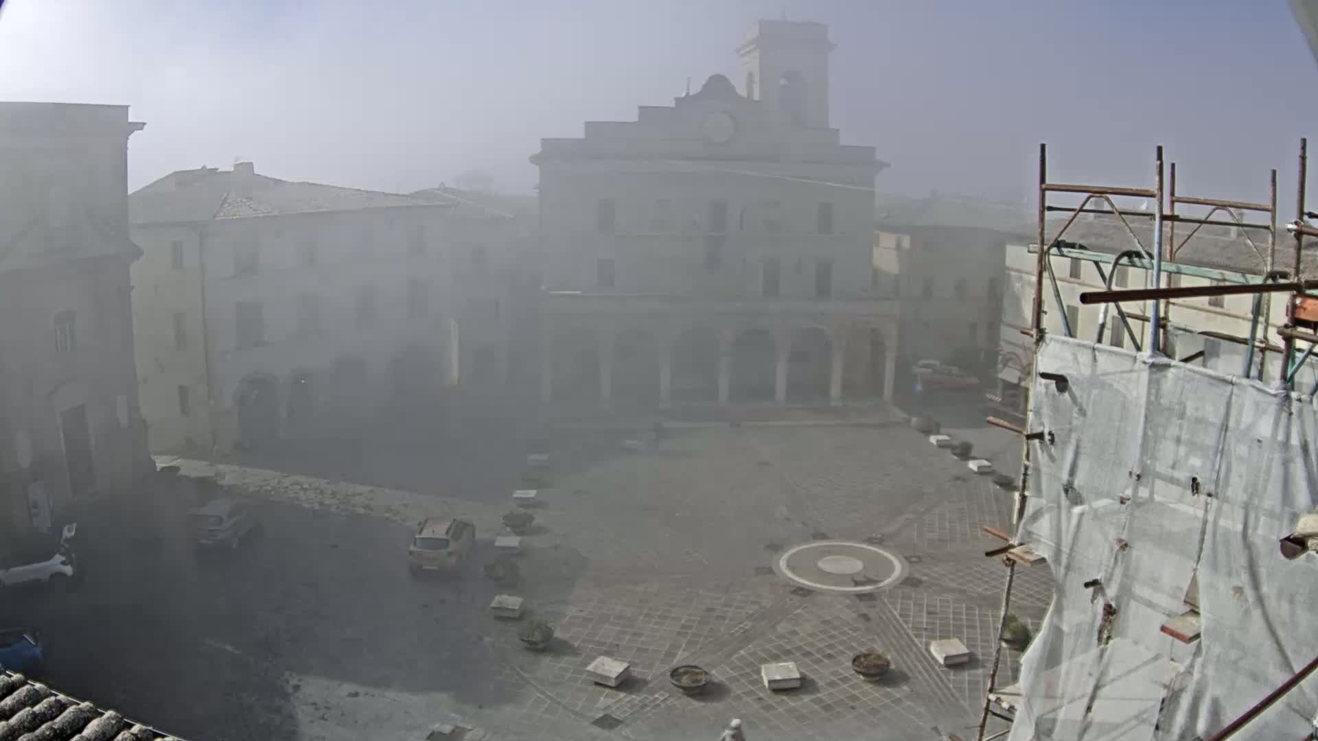 A foggy town square is seen with several buildings surrounding it, including a large church-like structure, and scaffolding with a tarp is in the bottom right corner.