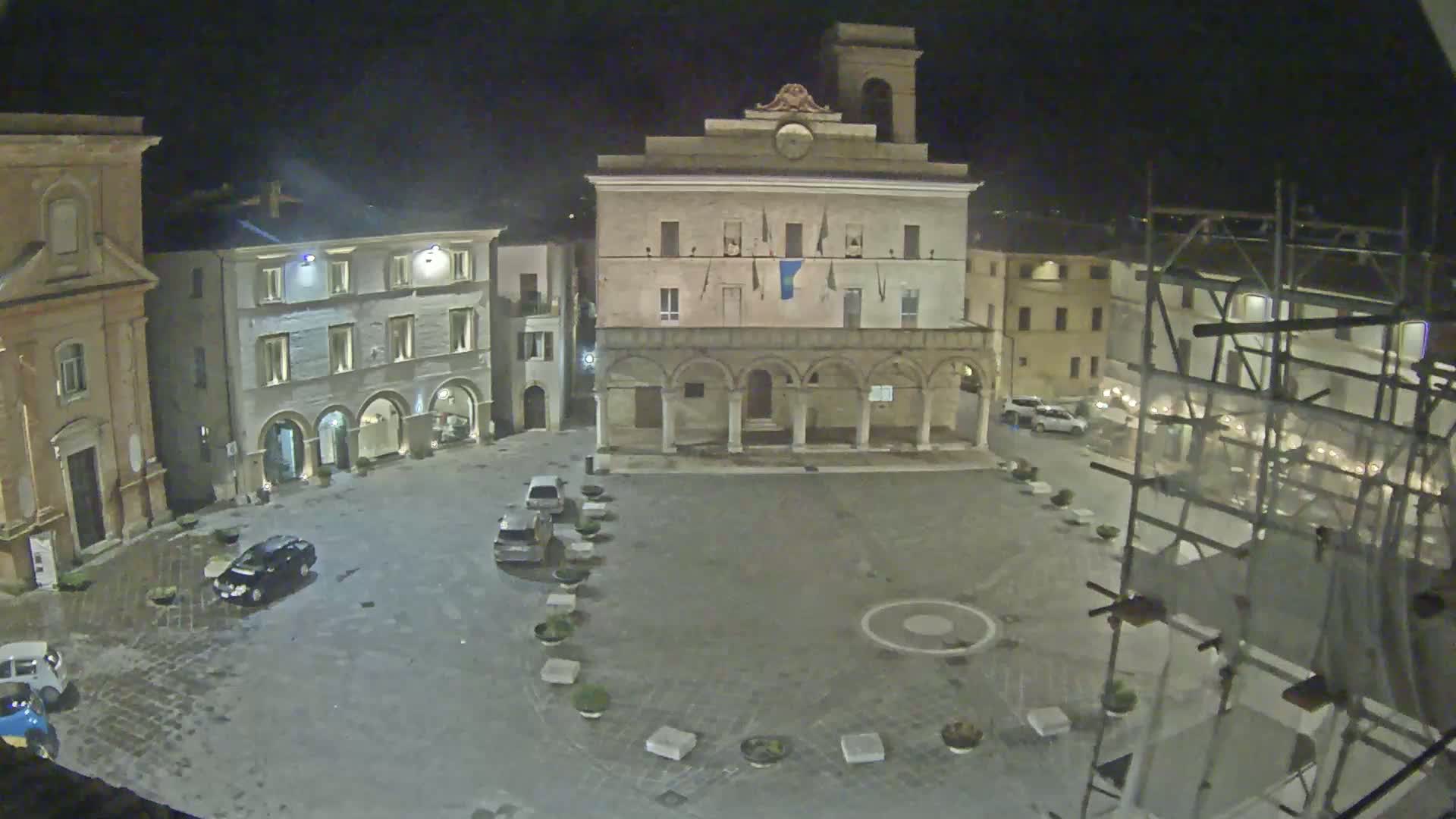 A nighttime view of a town square with several buildings, some cars parked, and scaffolding on one side, under slightly overcast conditions.