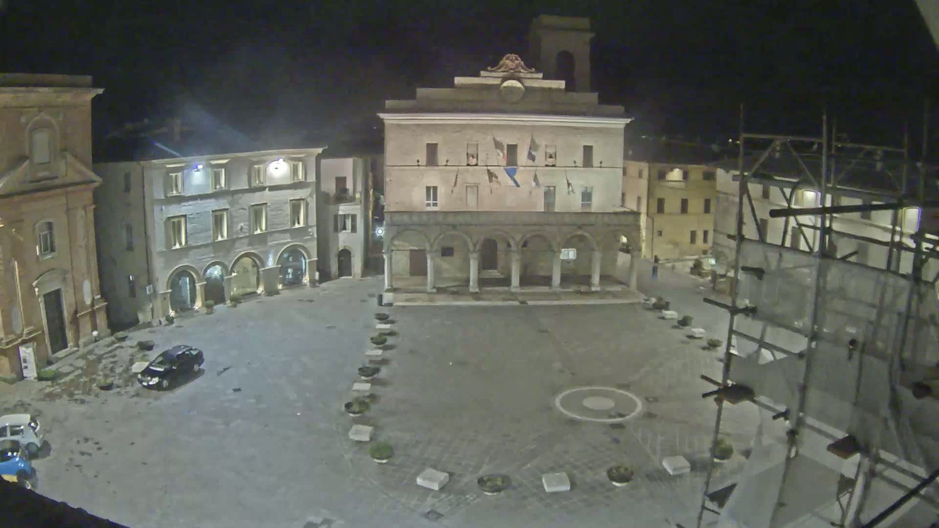 An empty town square at night, with several buildings surrounding it, one of which has scaffolding, under cloudy conditions.