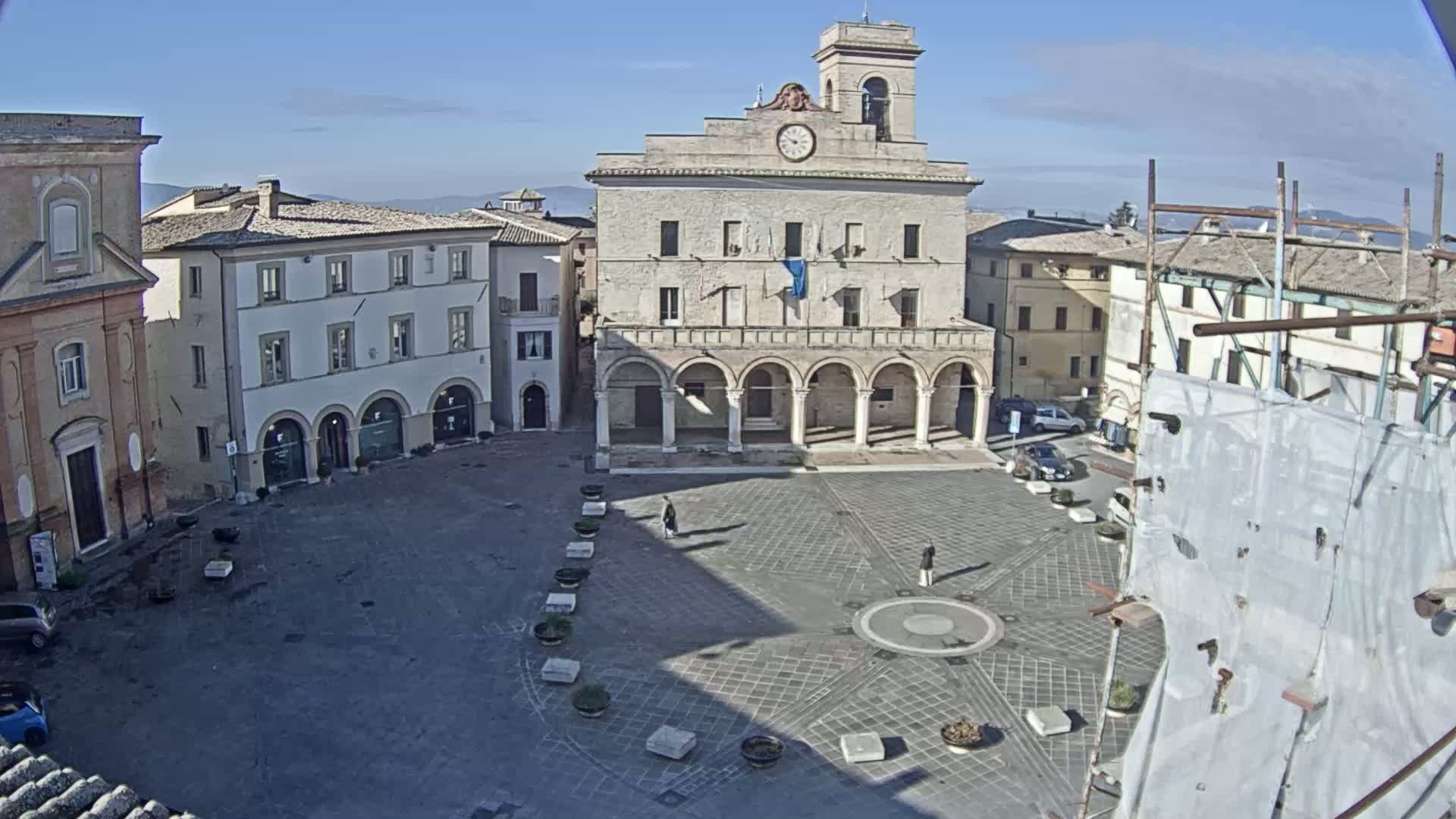 A mostly empty town square with surrounding buildings, under a clear sunny sky.