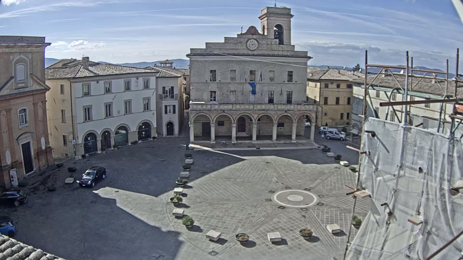 A paved town square, under a mostly sunny sky, is centered around a large building with arched entryways, surrounded by other buildings and a few parked cars.