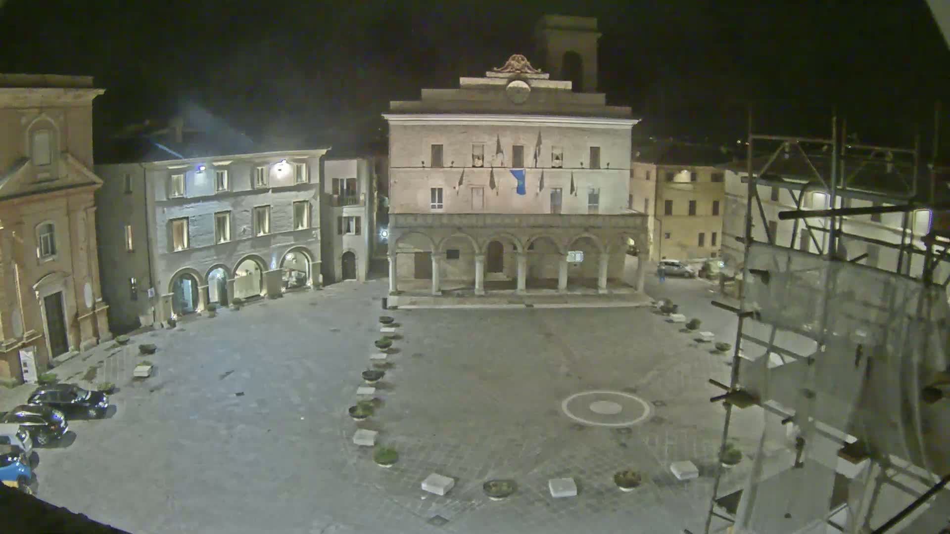 A town square at night, illuminated, features several buildings, including a large building with arches on the ground floor, some parked cars, and scaffolding on one side.