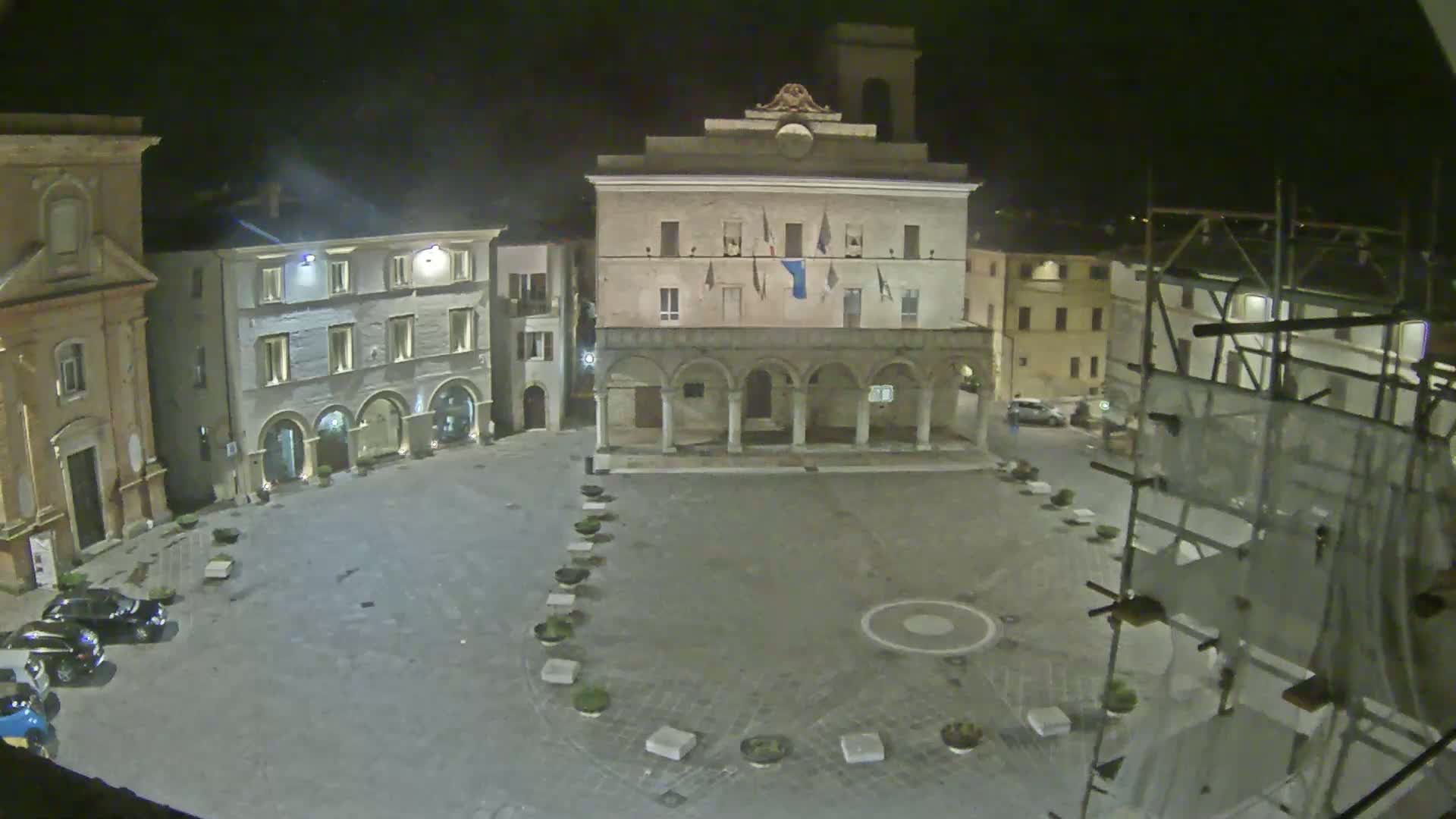 An empty, stone town square at night is illuminated, centered on a large building with arched columns and a few cars parked along the edge.