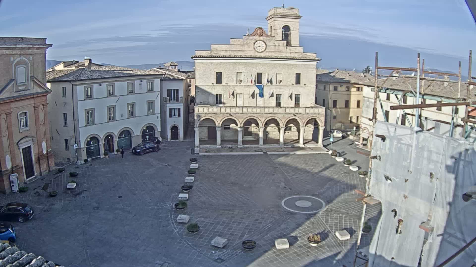 A town square, under a clear, sunny sky, is centered around a large building with arched entryways, surrounded by other buildings and a few cars.