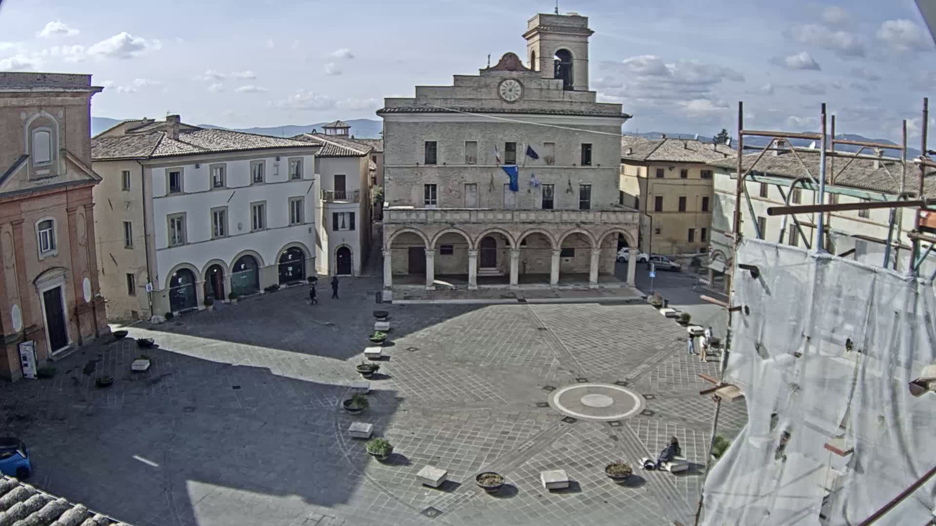 A mostly empty town square, under a partly cloudy sky, features a large stone building with arched entryways, surrounded by other buildings.