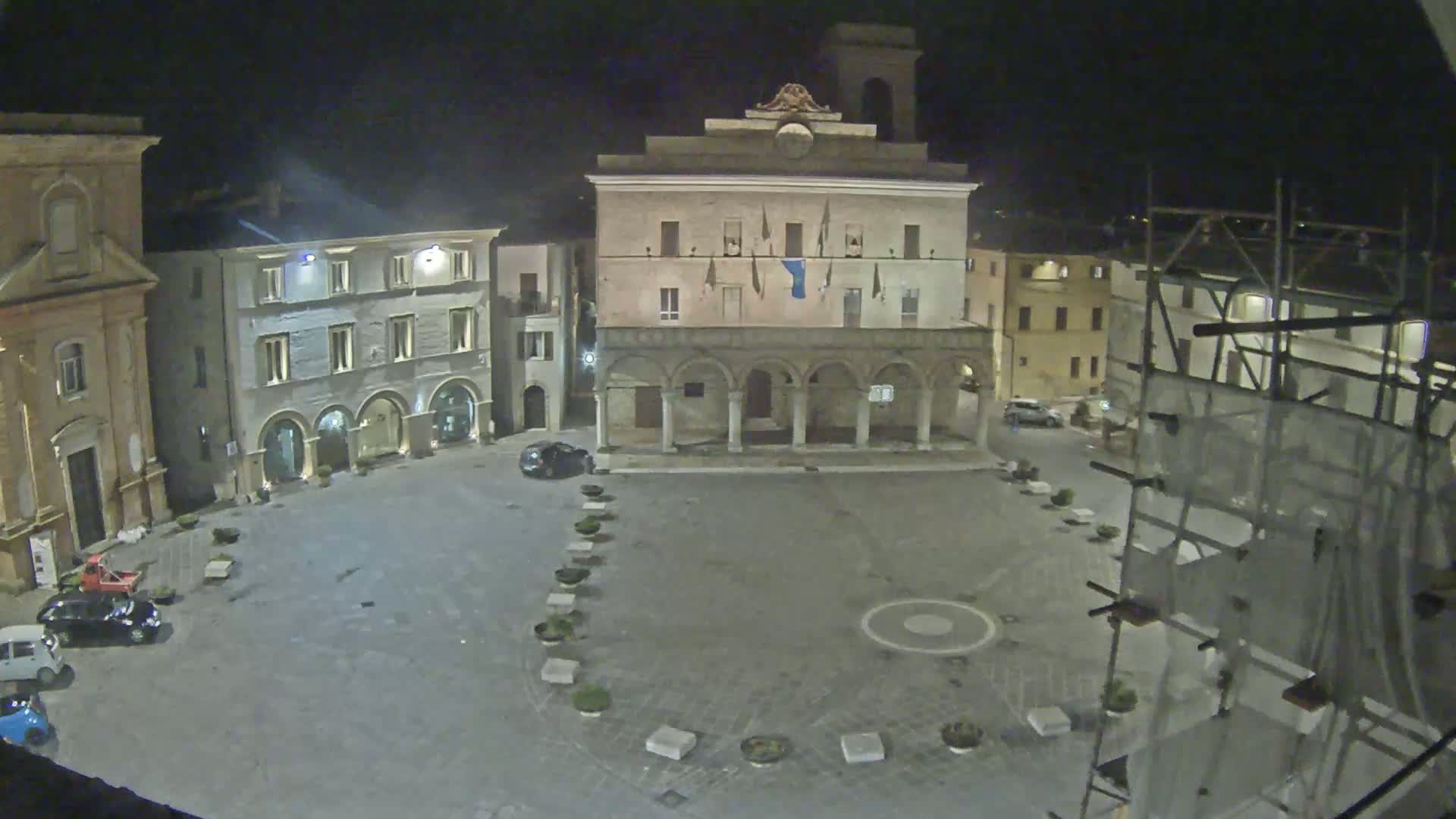 A nighttime view of a town square with several buildings, including a large, arched building in the center, and a few cars parked, under a dark sky.