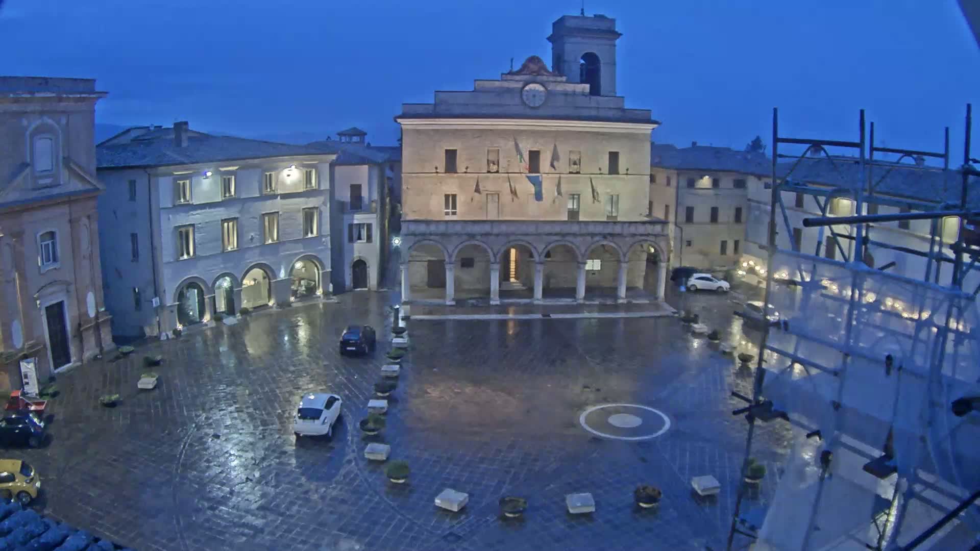 A town square at night, under a dark, rainy sky, is centered around a large building with arched entryways, surrounded by smaller buildings, a few cars, and construction scaffolding.