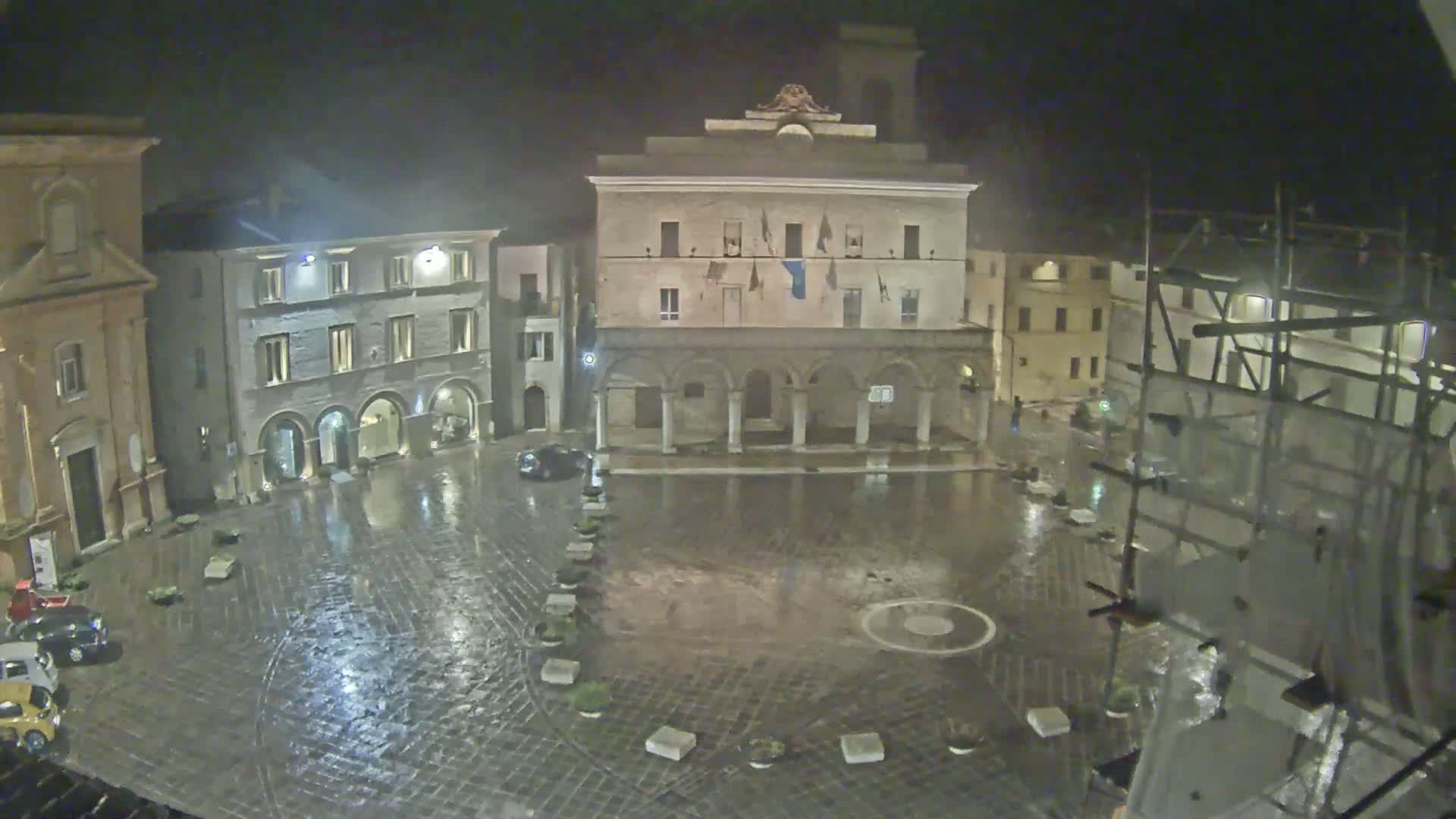 A rain-slicked town square at night, surrounded by historic buildings, one of which has an arcade.