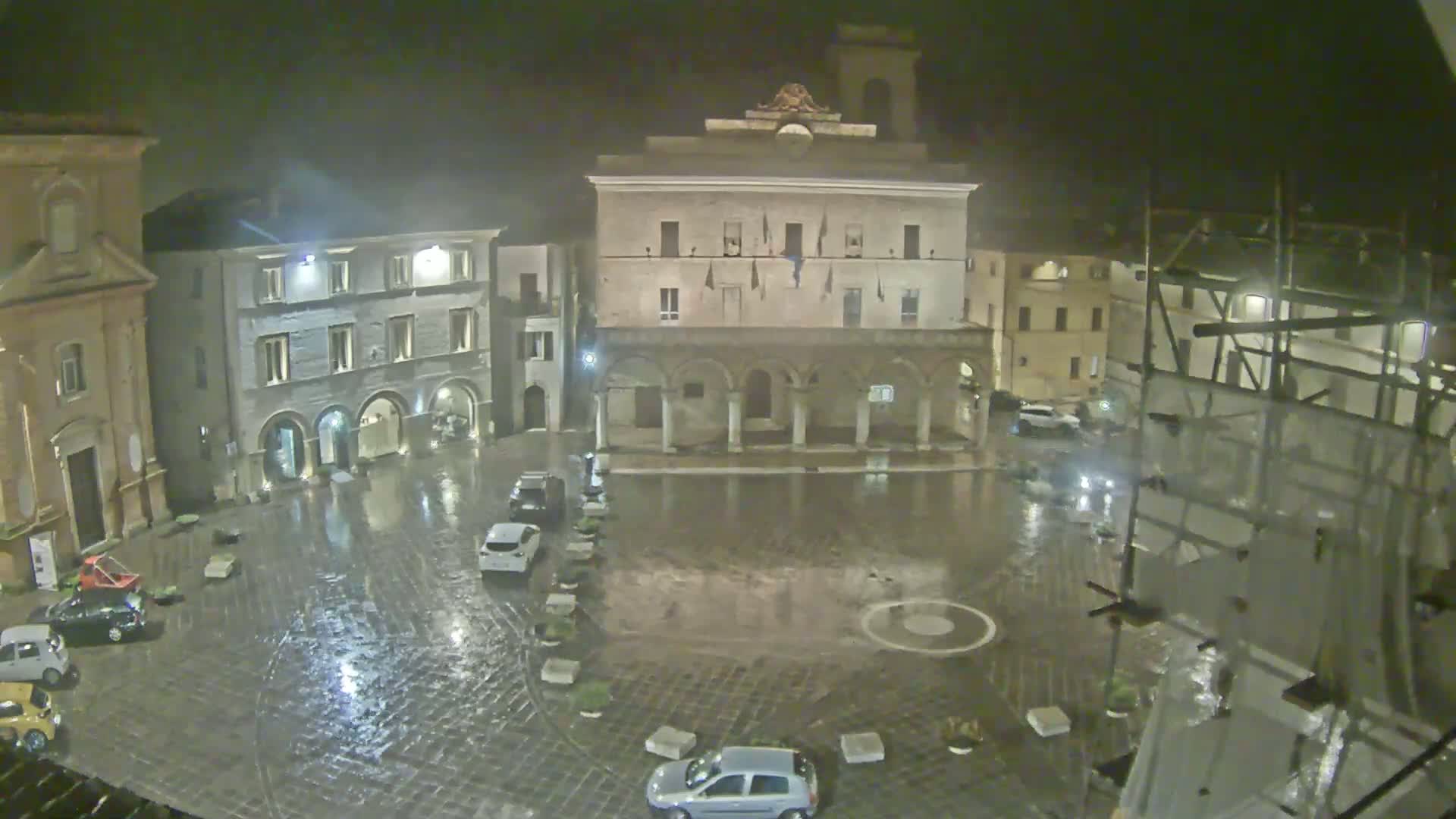 A rain-soaked town square at night is surrounded by historic buildings, some under construction, with a few cars parked around the perimeter.