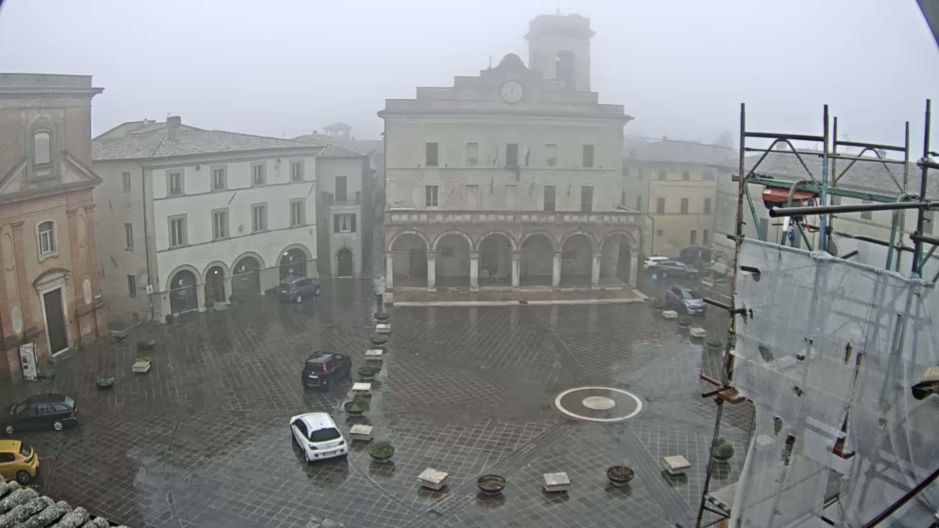 A wet, foggy town square is seen with several cars parked and buildings surrounding it, one featuring an arched colonnade and a clock tower.
