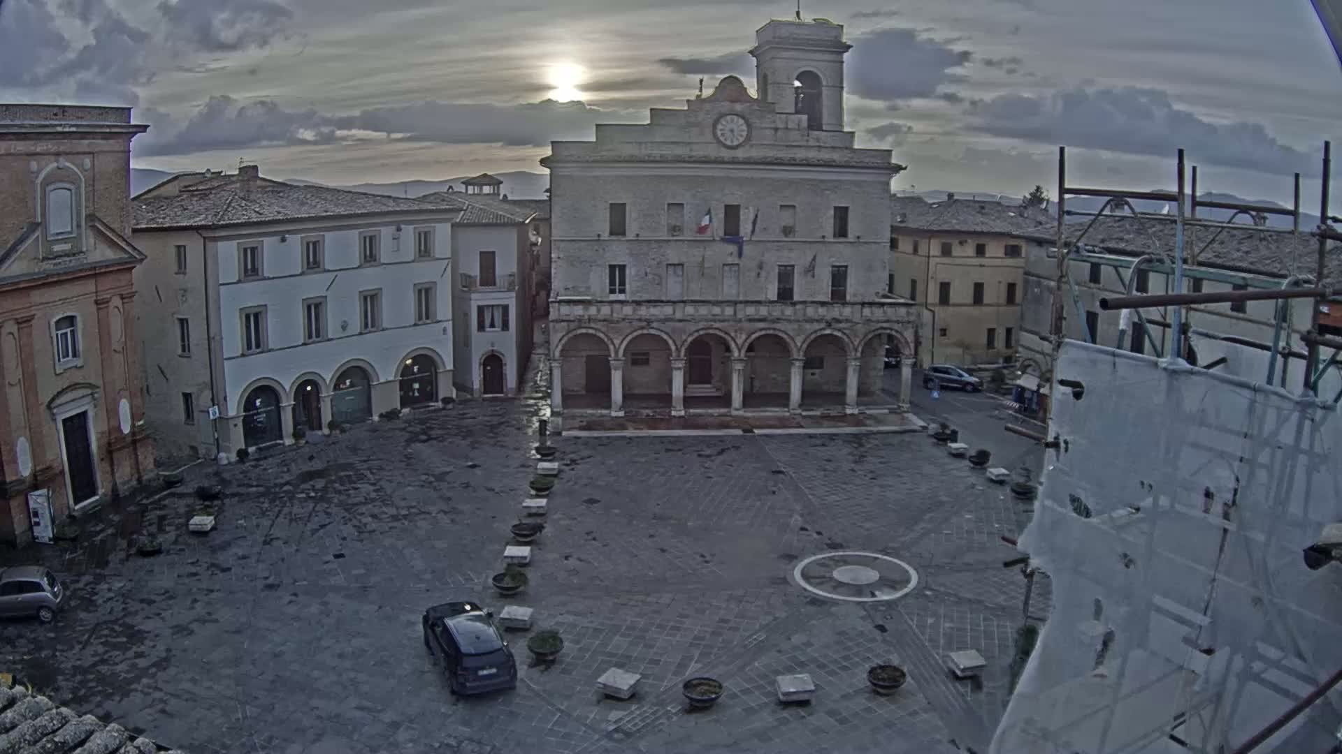 A town square with a large building featuring arched entryways, surrounded by other buildings, under an overcast sky.