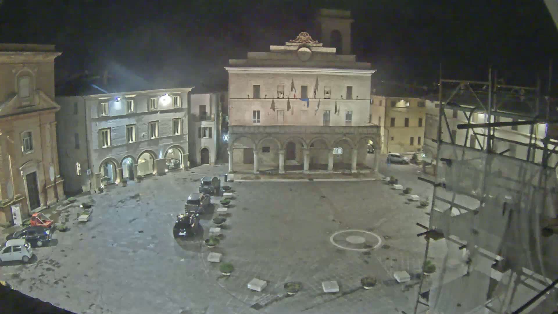 A nighttime view of a town square with several buildings, some cars parked along the side, and scaffolding on one building.