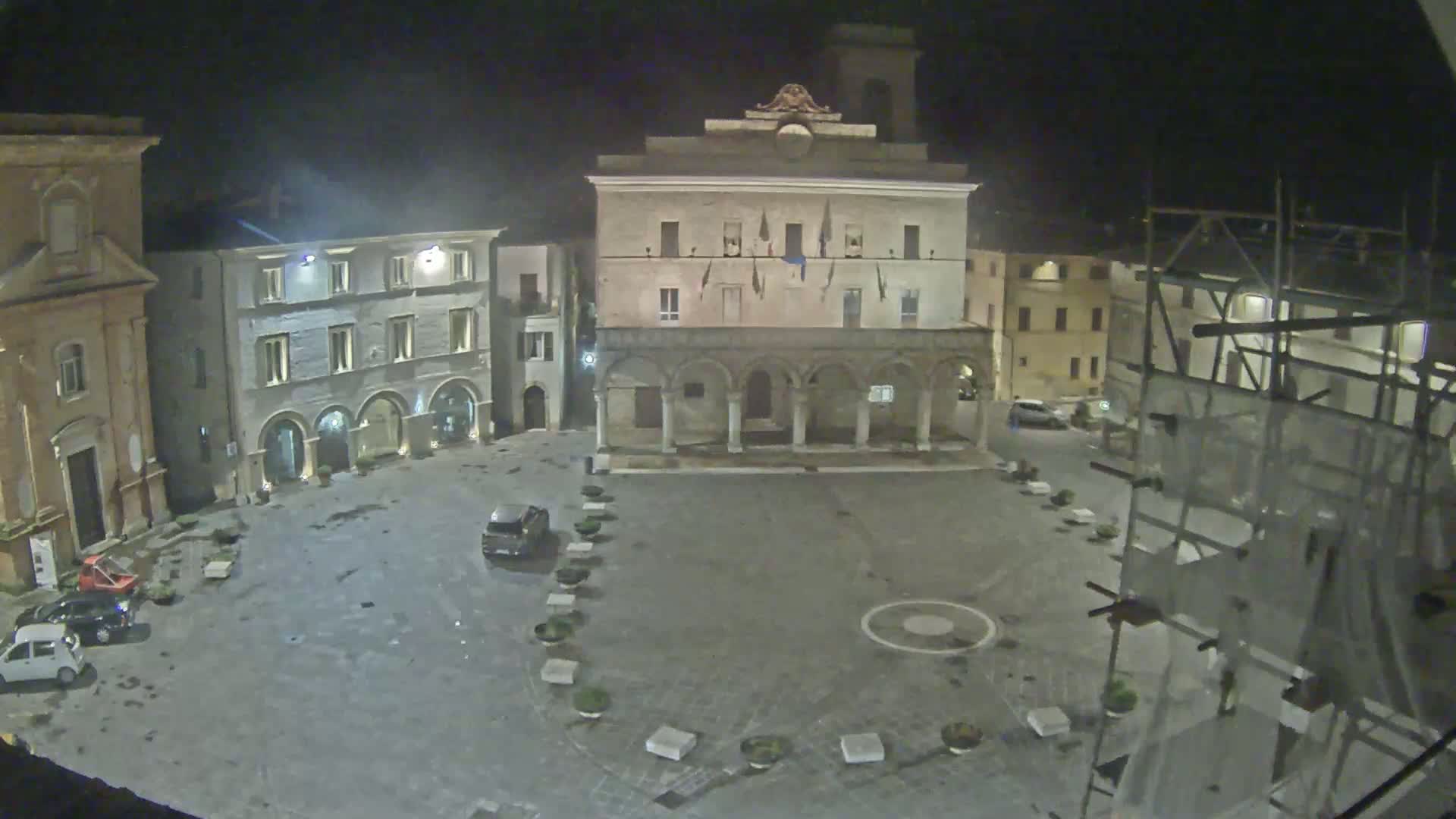 A nighttime, mostly empty town square is seen, with several buildings surrounding it, including a large stone structure with arched entryways, under dark, possibly overcast skies.