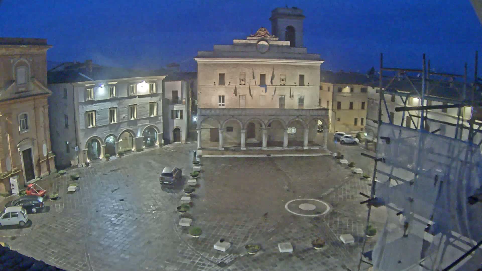 A town square at night, under a dark blue sky, features several buildings including a large stone structure with arched entryways and a clock tower, surrounded by parked cars.