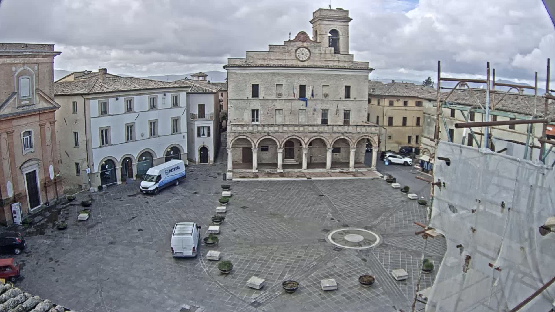 A town square with a large stone building, several smaller buildings, and a few parked vehicles is visible under cloudy skies.