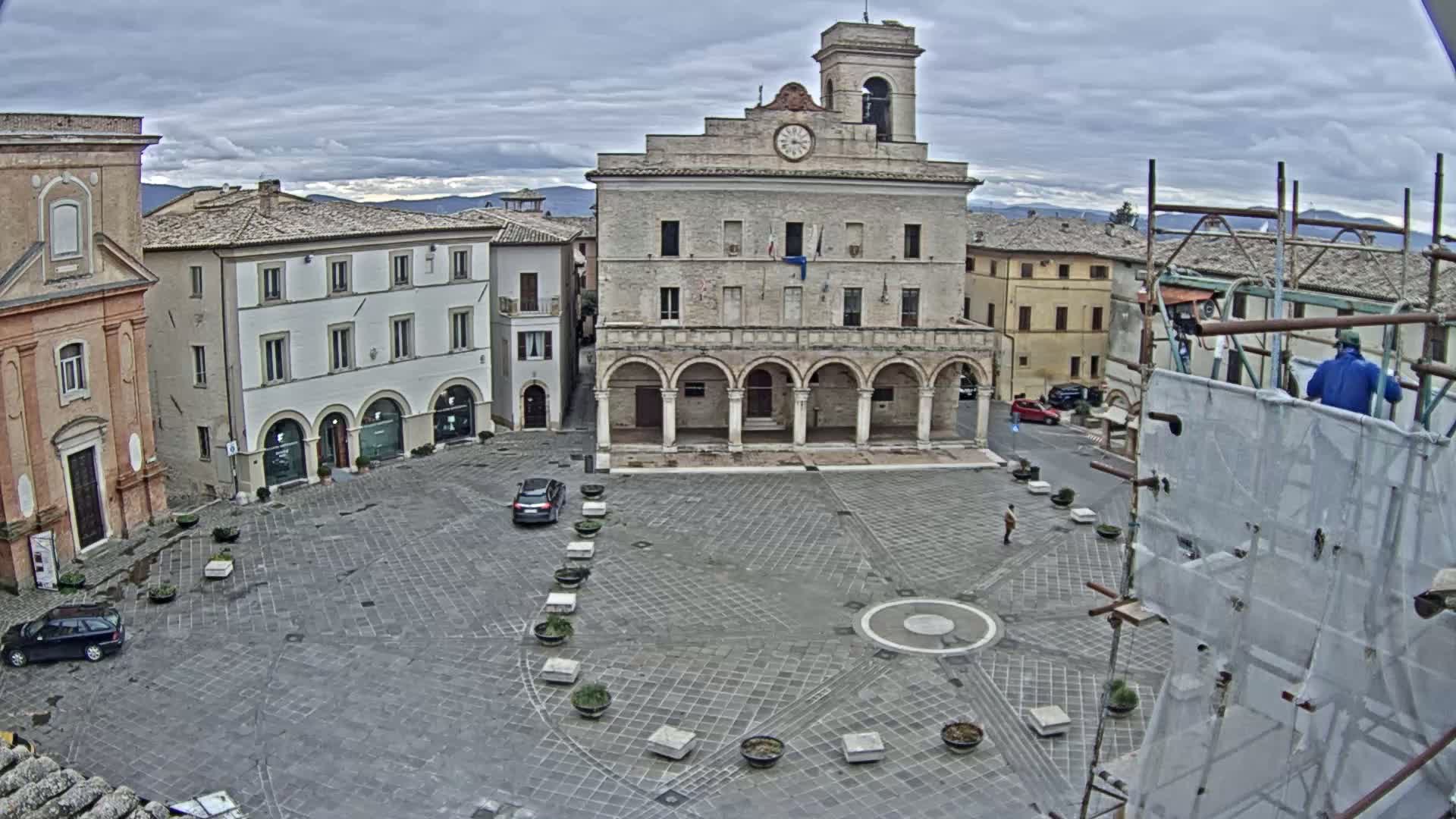 A town square with a large stone building, several smaller buildings, a few cars, and scaffolding under an overcast sky.