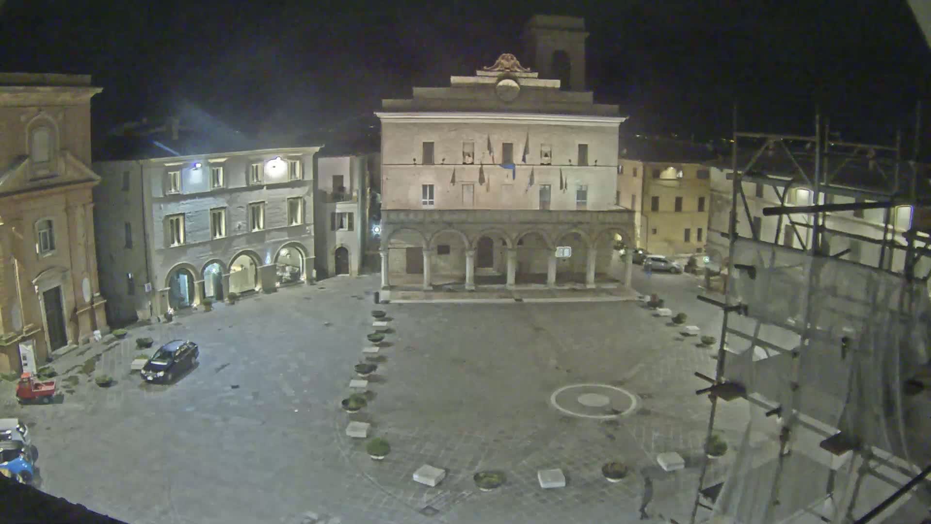 A nighttime view of a town square, featuring several buildings, one with a colonnade, and scaffolding on a building to the right, under dark conditions.