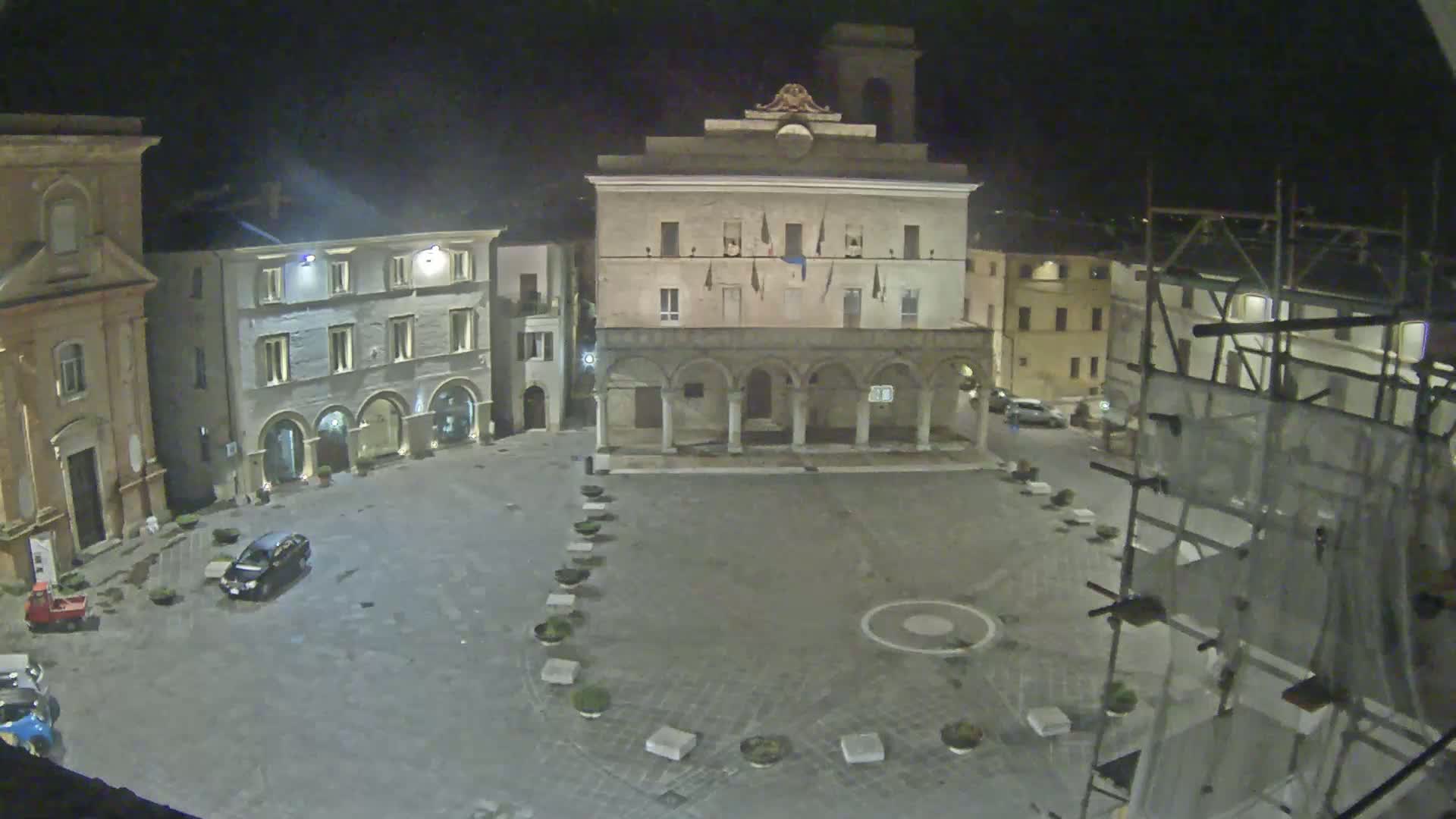 A nighttime view of a town square, featuring several buildings, a few cars, and scaffolding on one building.