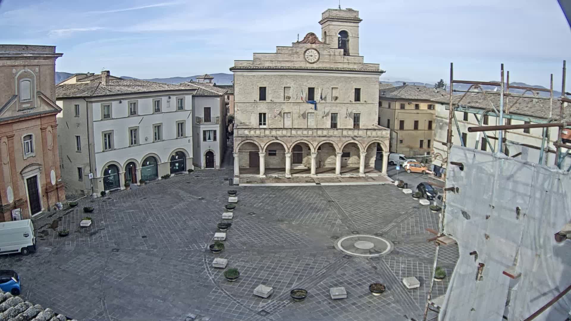 A mostly empty cobblestone town square is centered on a large building with a clock tower under a partly cloudy sky.