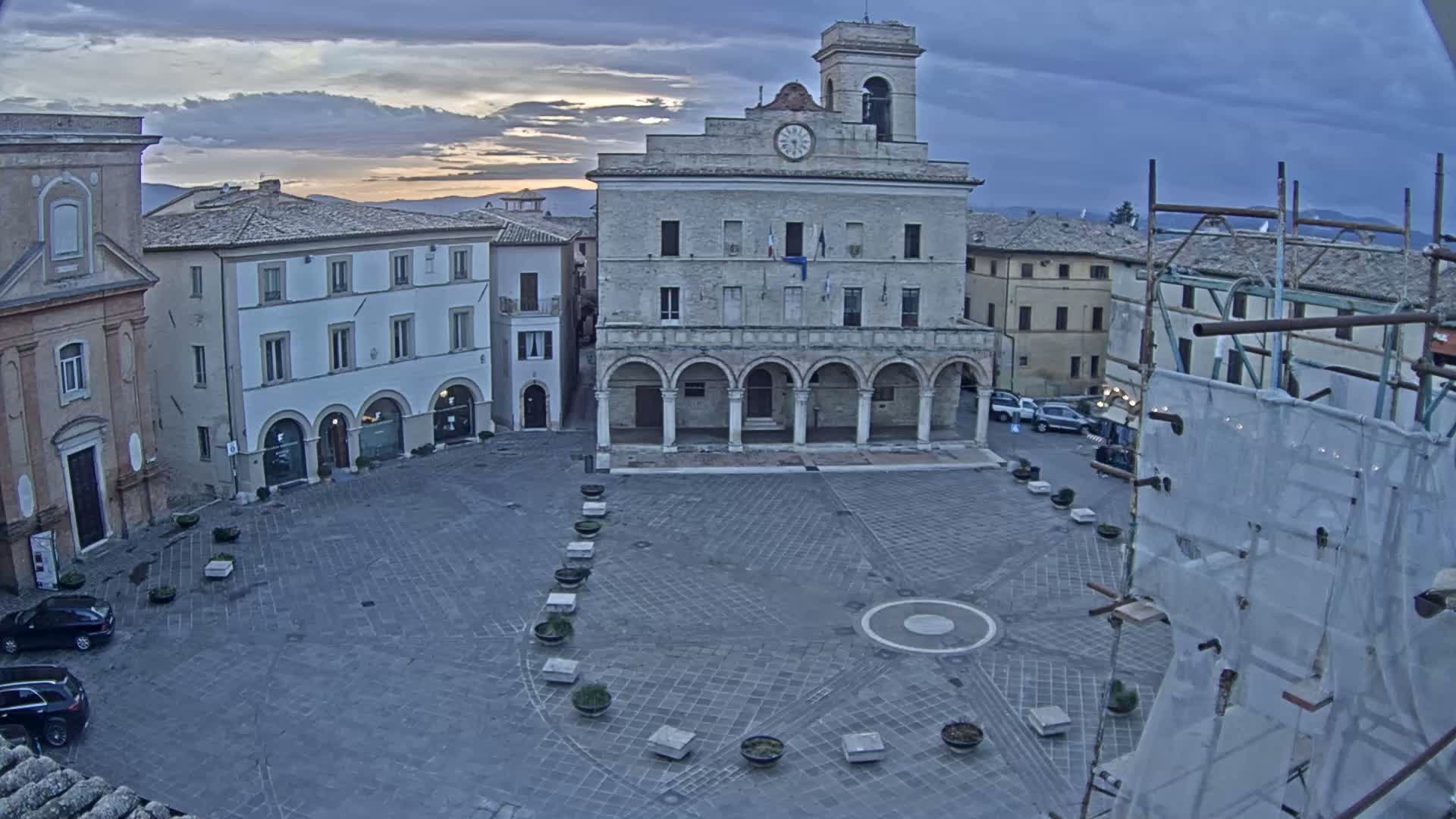 A town square with a large building featuring arched entryways and a clock tower is visible under an overcast sky at sunrise.