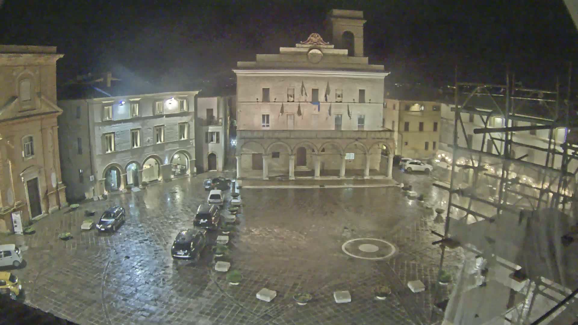 A rain-slicked town square at night, surrounded by historic buildings, with several cars parked along the edge.