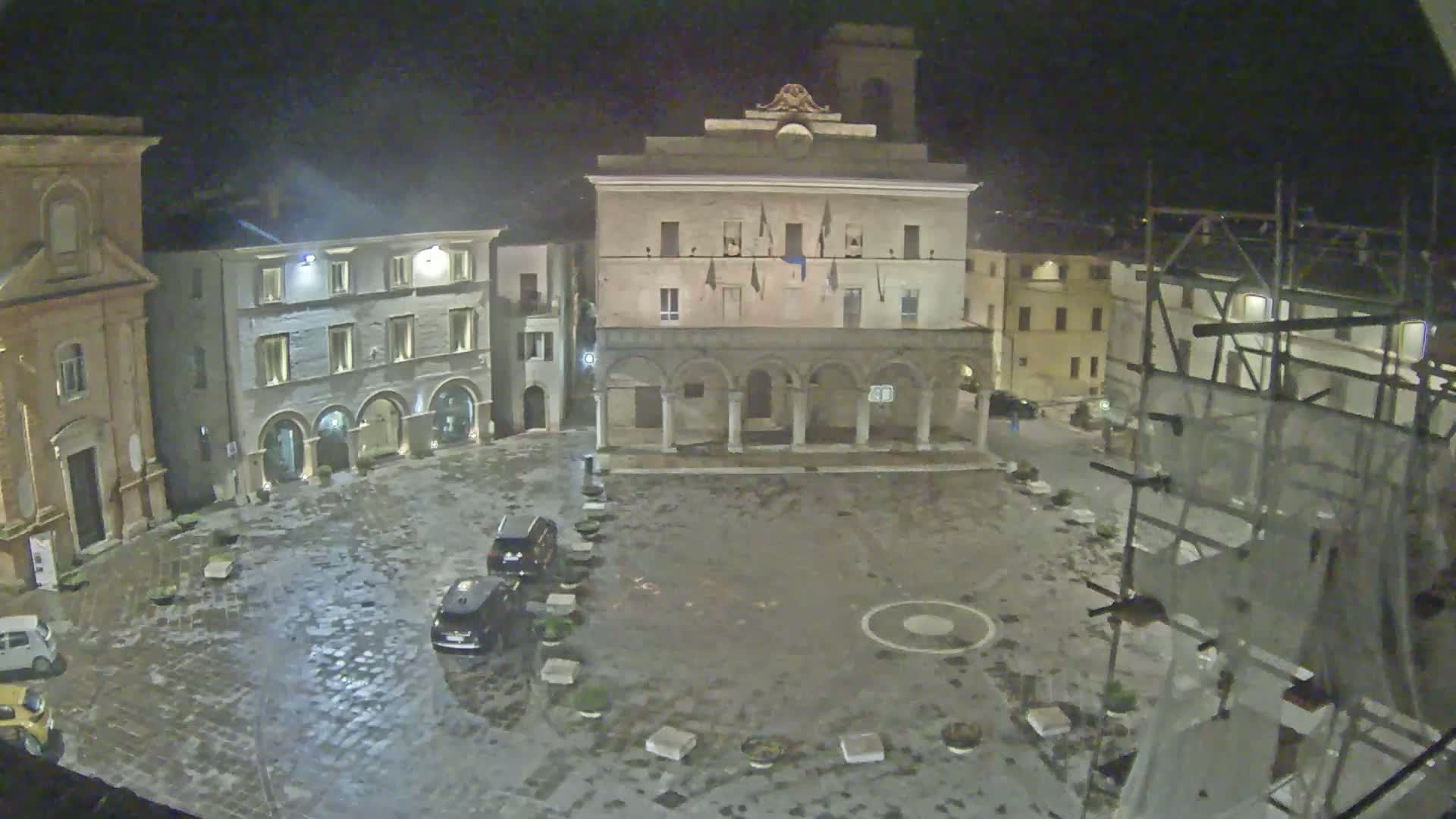 A wet, cobblestone town square at night is surrounded by historic buildings, one of which is under construction.