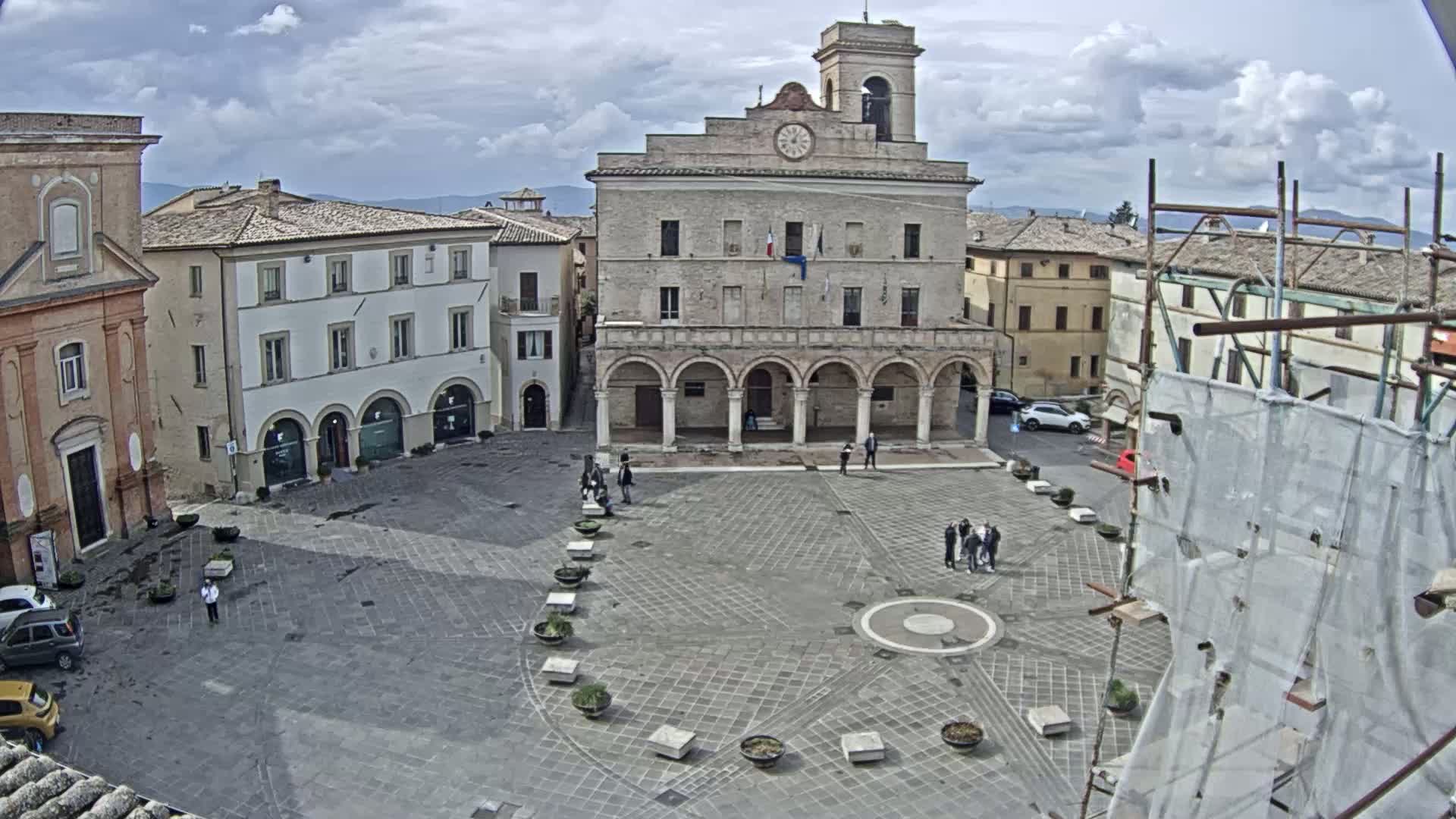 A large town square with a clock tower and several buildings is visible under a partly cloudy sky.