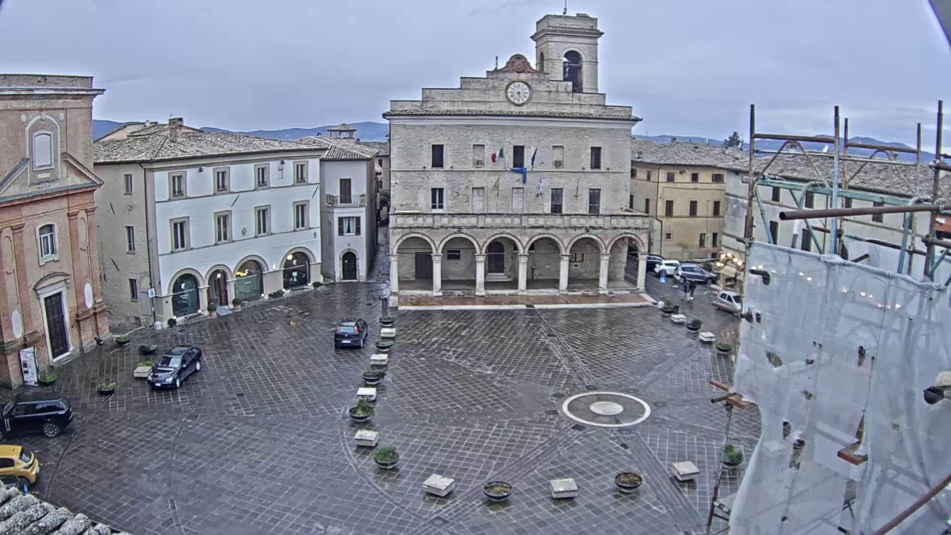 A large, stone plaza with several buildings surrounding it, including a large building with arched entryways and a clock tower, under an overcast sky.