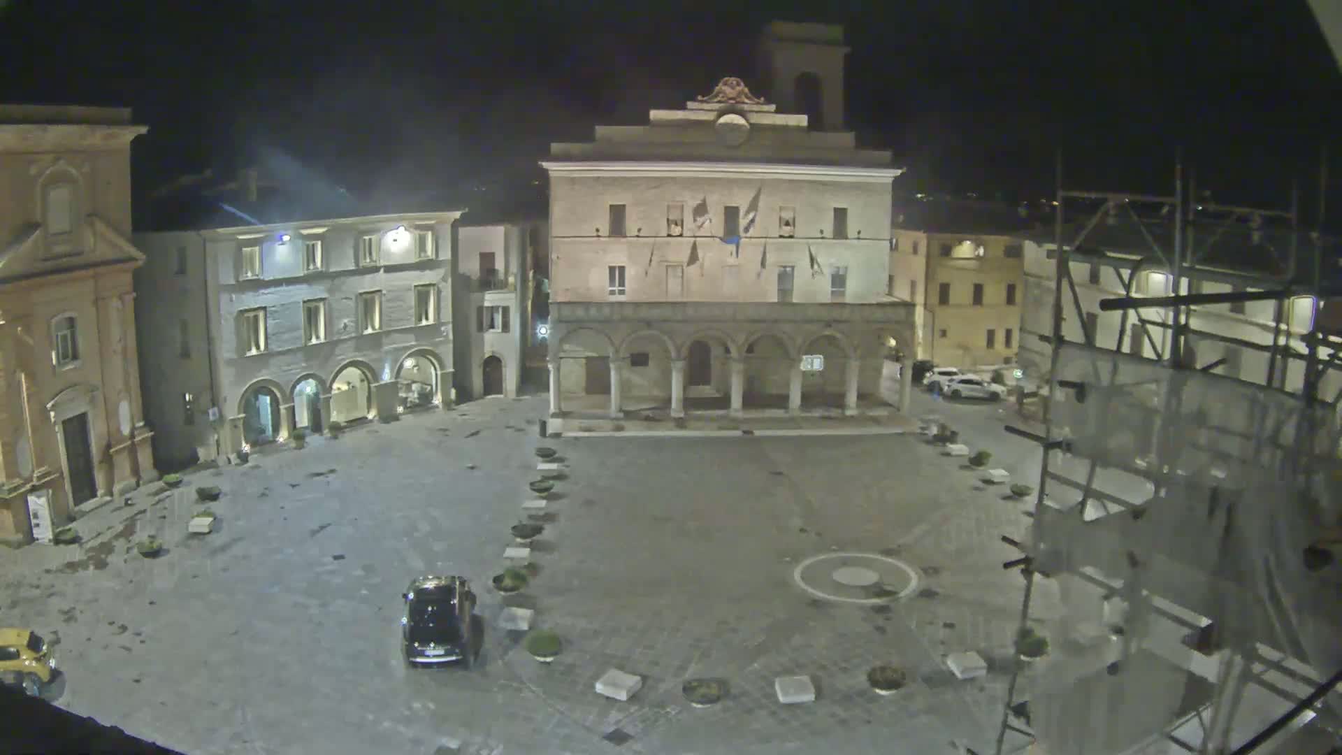 An empty town square at night, illuminated by artificial light, features several buildings, including a large stone structure with arches, and scaffolding surrounding a building under construction.