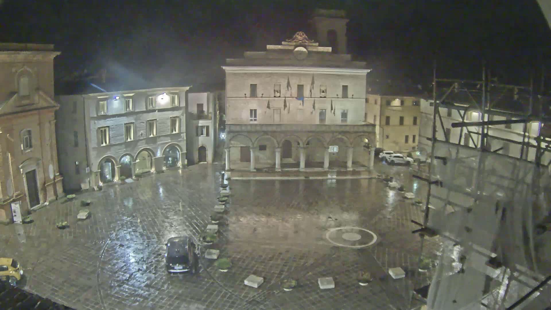 A rain-soaked town square at night, surrounded by historic buildings, with scaffolding visible on one side.
