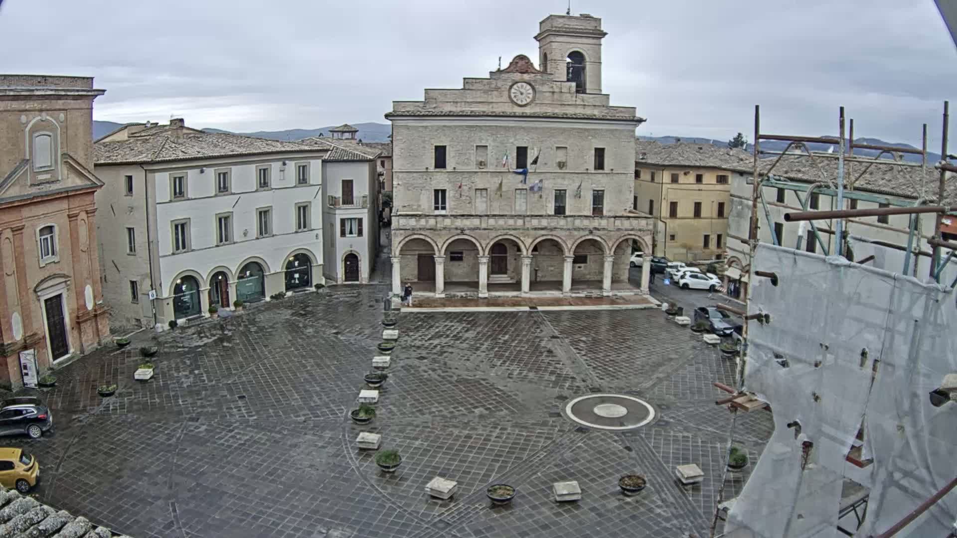 A large, stone town square, under an overcast sky, features a prominent building with arched entryways and a clock tower, surrounded by other buildings and a few parked cars.