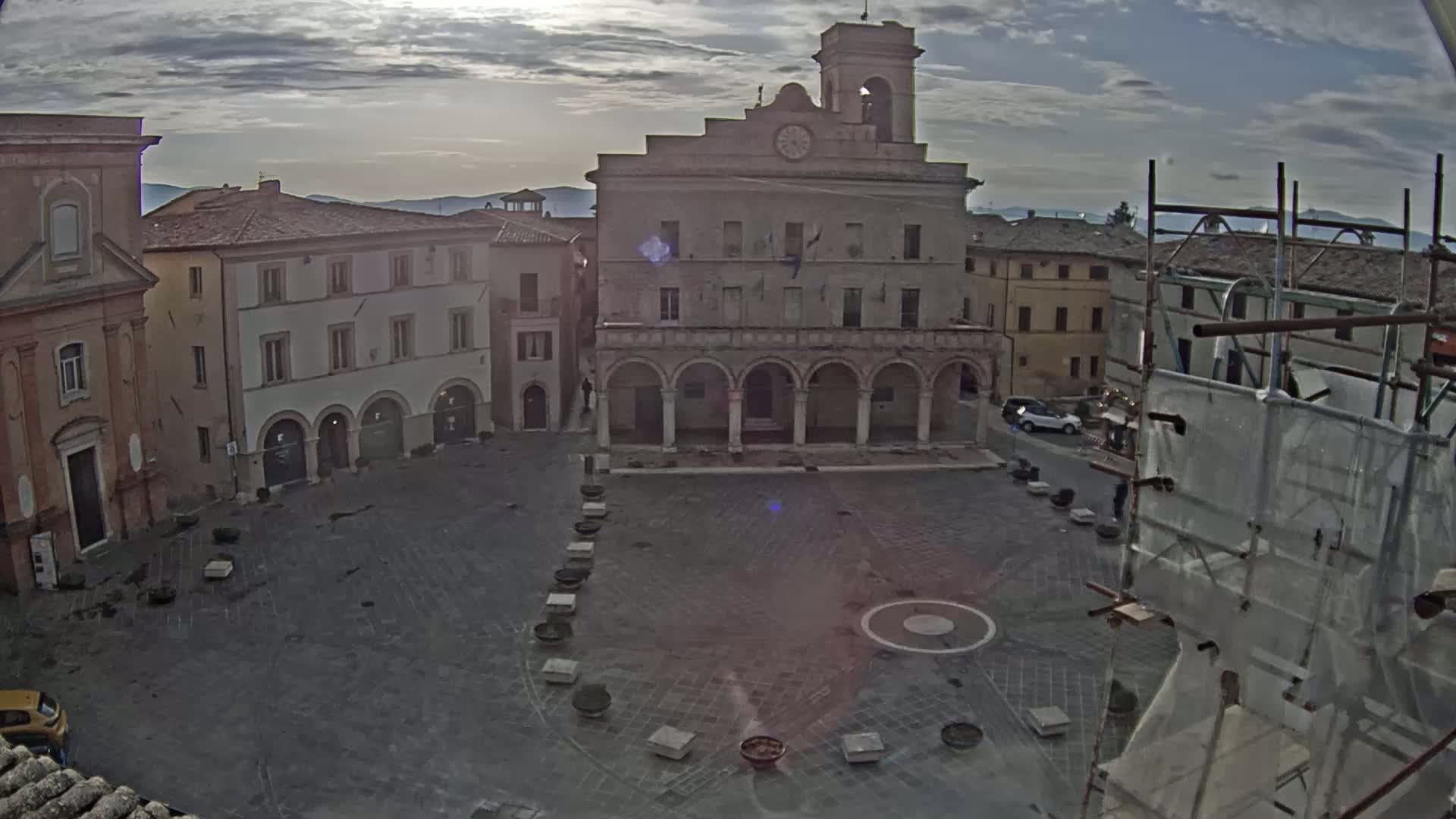 A mostly empty town square, under a partly cloudy sky, features a large stone building with arches and a clock tower, surrounded by other buildings.