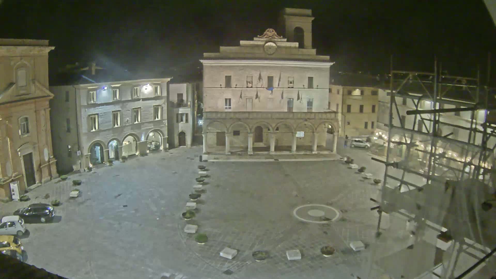 A nighttime view of a town square, featuring several stone buildings, a large central building with arches, and scaffolding on one side.