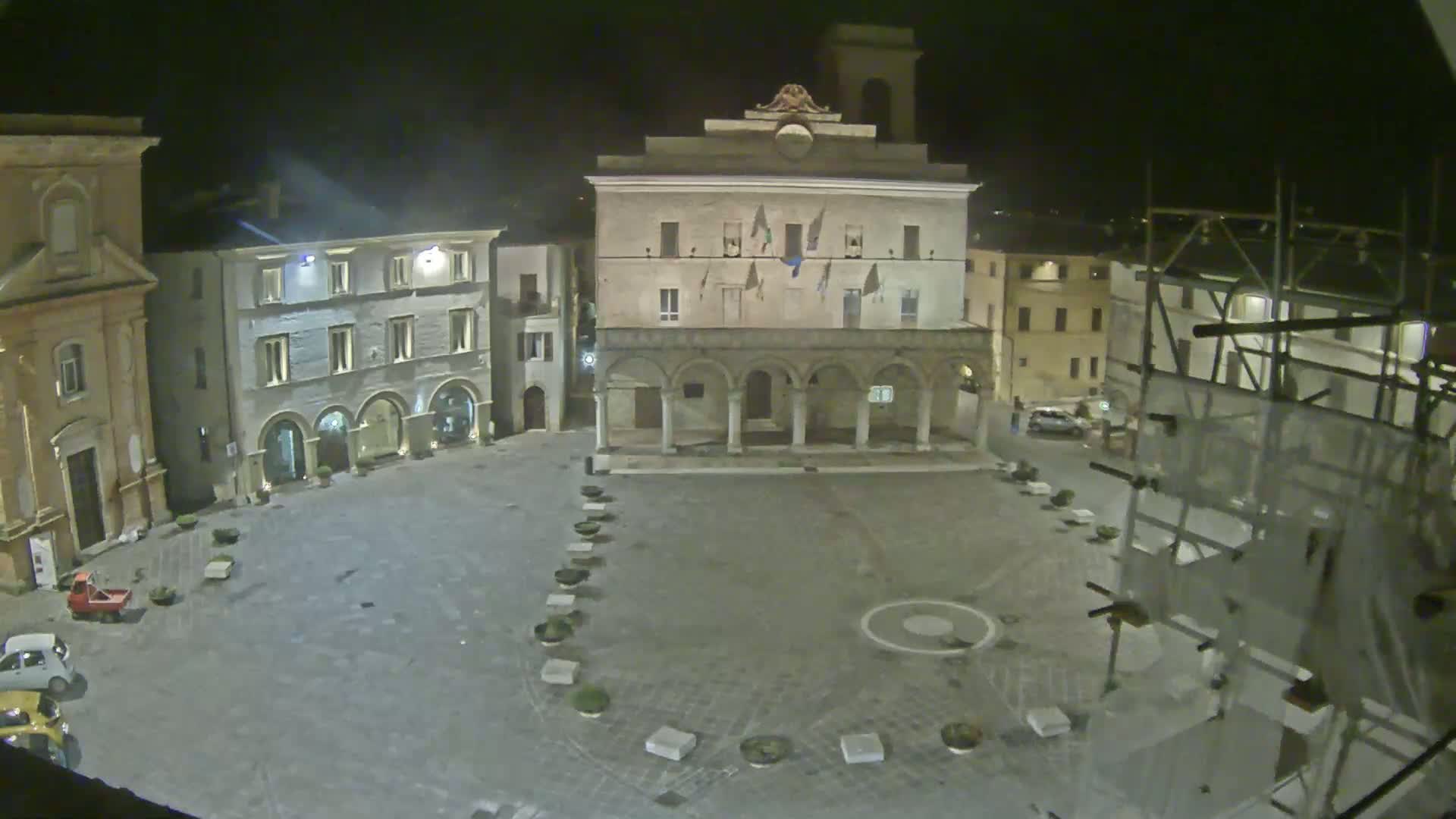 A nighttime view of an empty town square, surrounded by stone buildings, with scaffolding visible on one side.