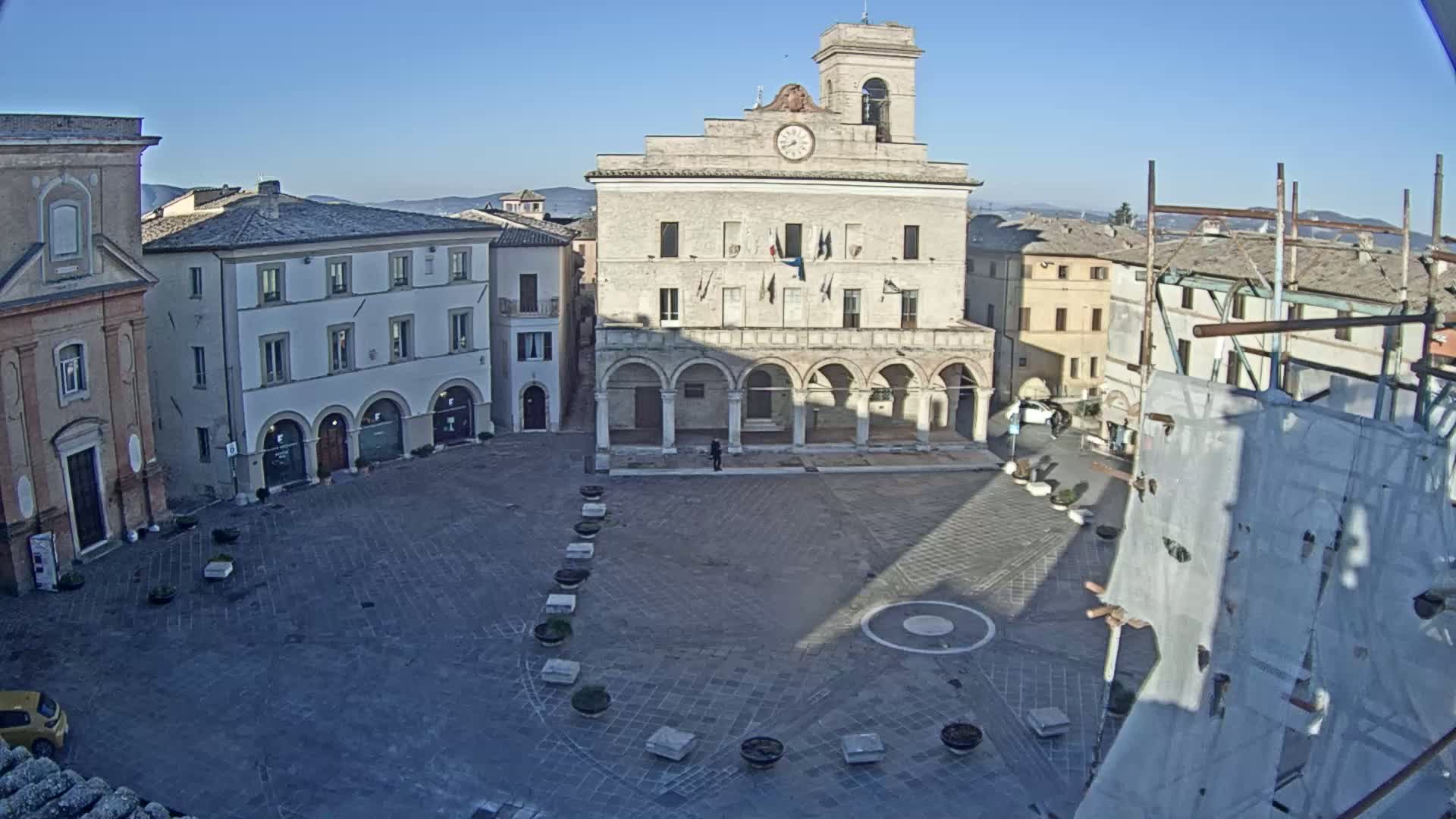 A mostly empty town square, under a clear blue sky, is centered around a large building with a clock tower and arched colonnade.