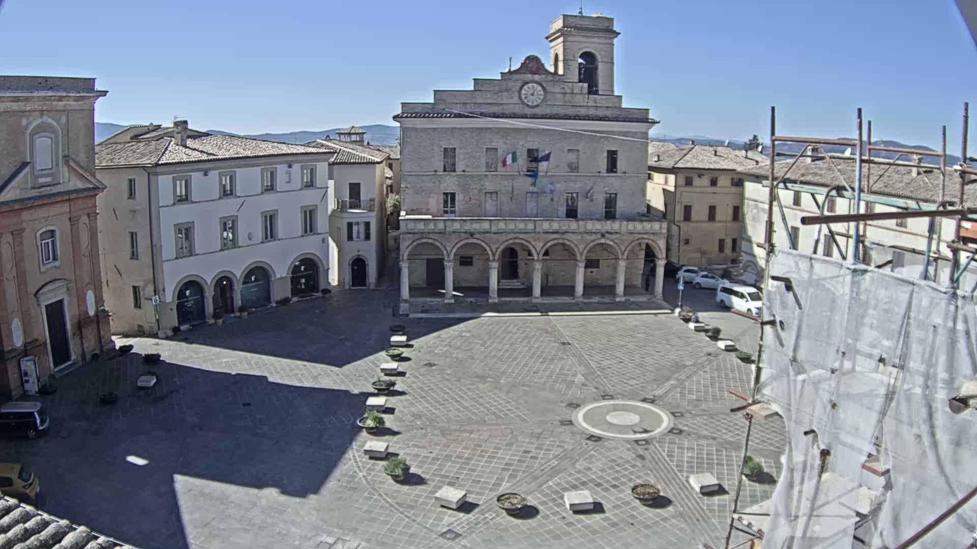 A large town square, paved in a geometric pattern, is centered on a large building with arched colonnades under a clear, sunny sky.